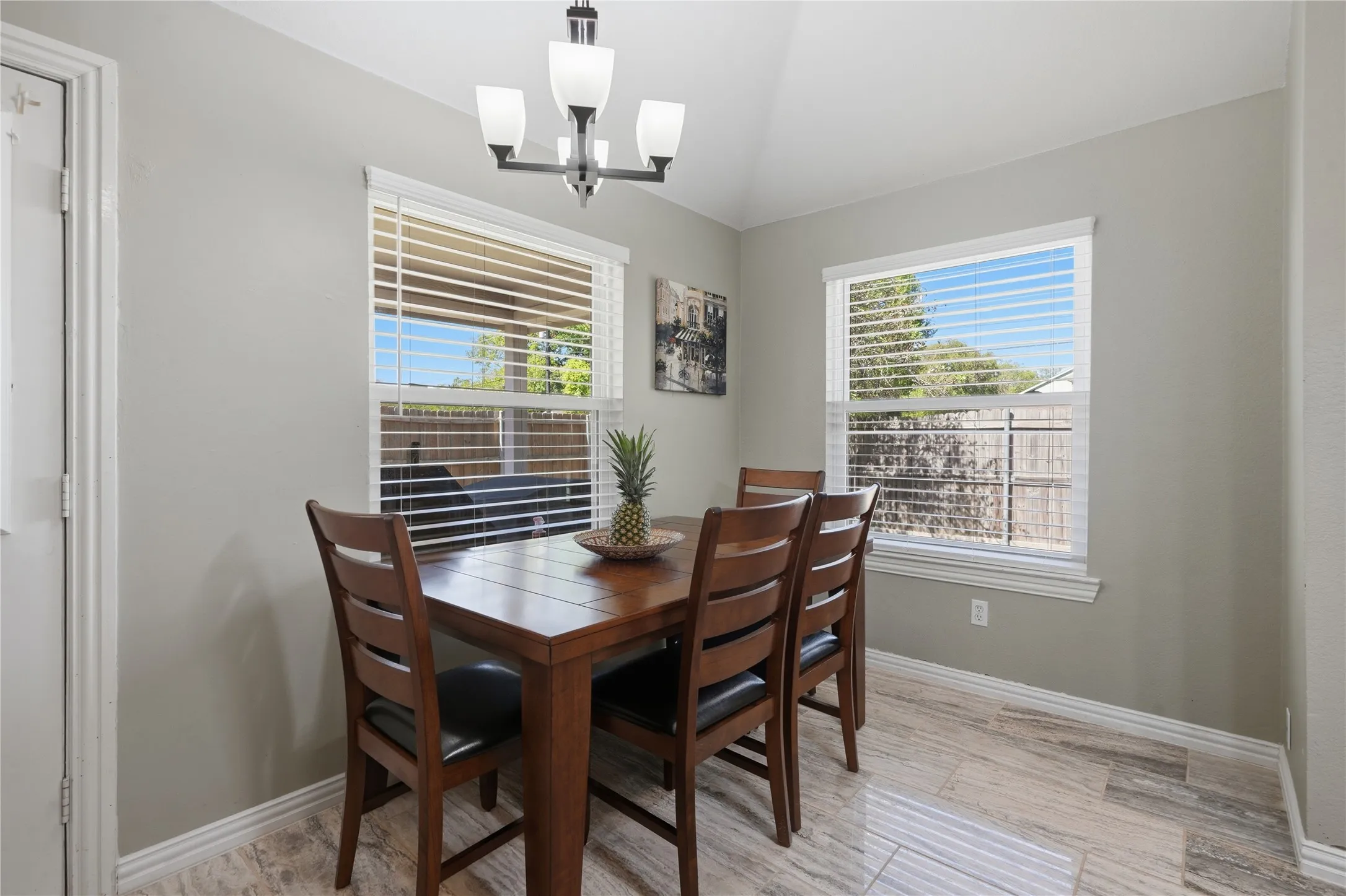 Vaulted ceiling, contemporary chandelier, two windows with blinds, light gray walls, and wood-finish tile flooring