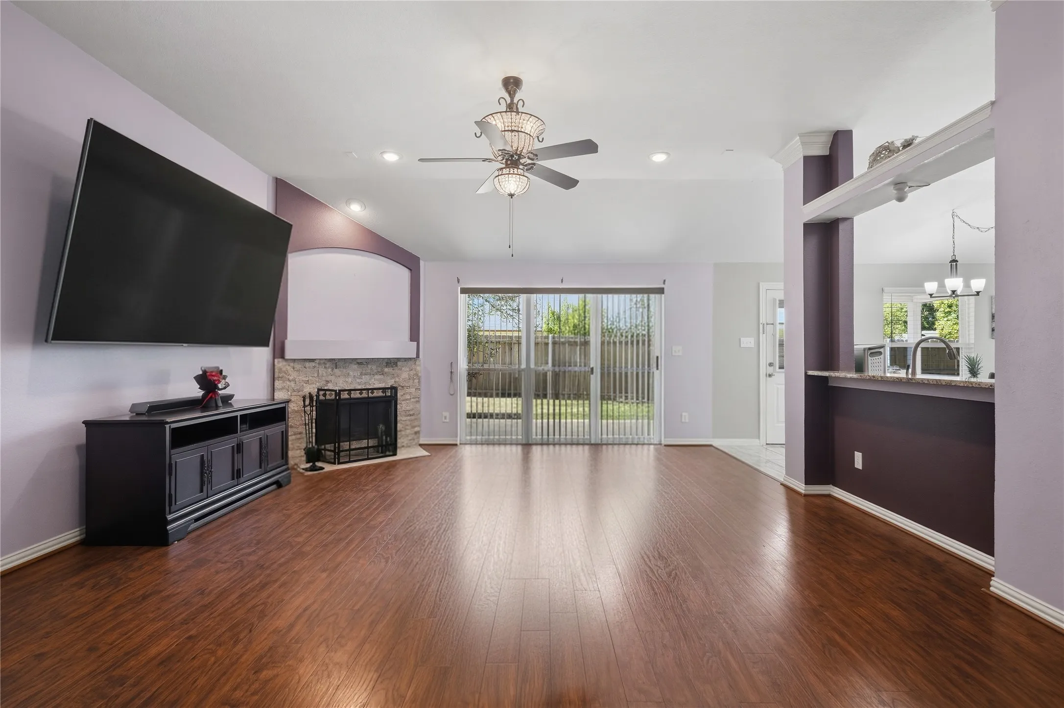 Spacious living area featuring wood-finish flooring, a stone-faced fireplace with hearth, and a ceiling fan with integrated lighting
