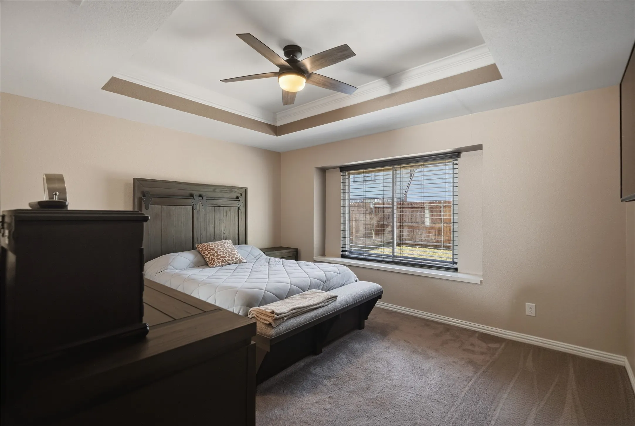 Recessed tray ceiling with crown molding, ceiling fan with integrated lighting, window with blinds, and carpeted flooring