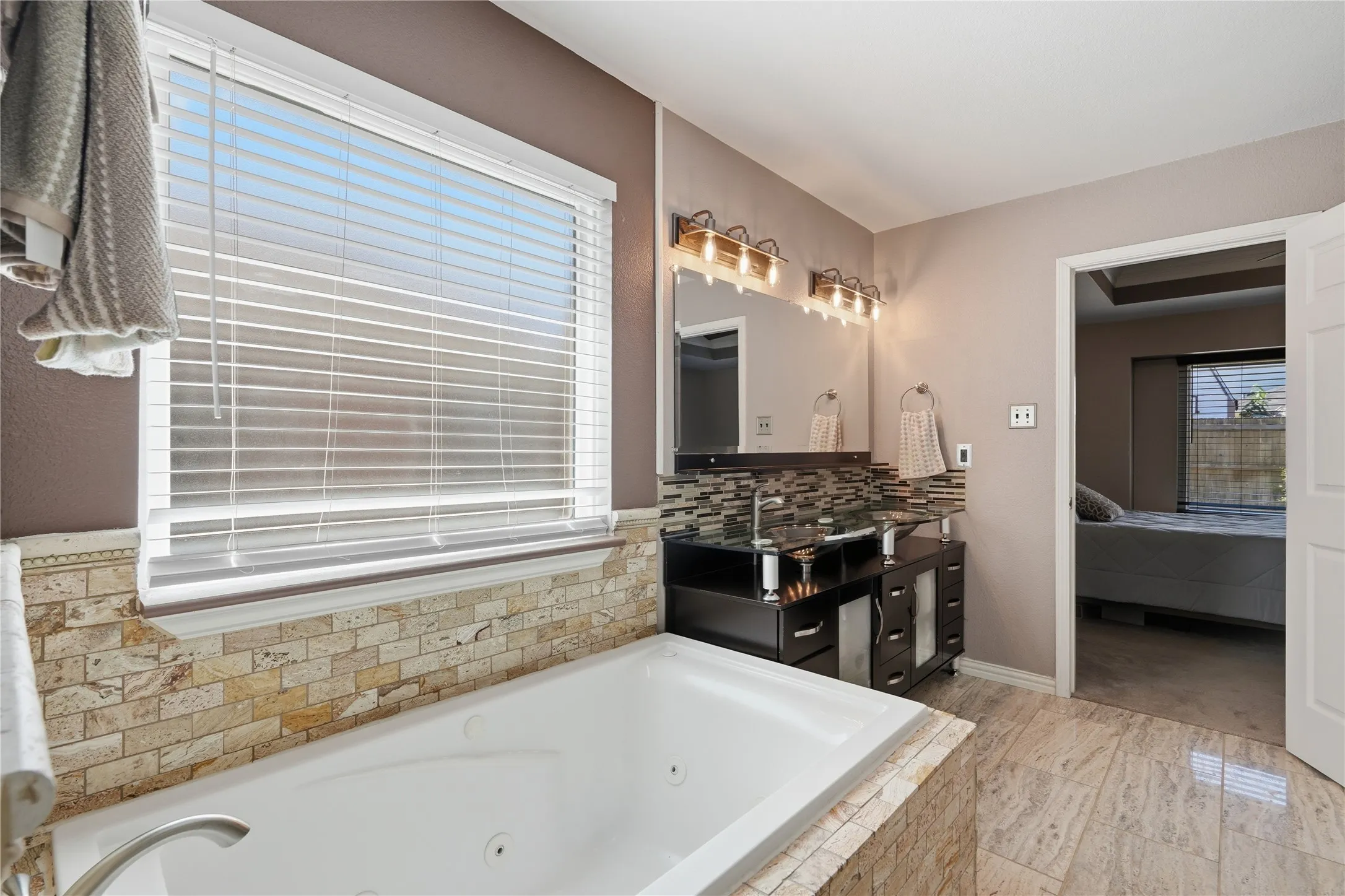 Jetted soaking tub with stone tile surround, dual vanity with dark wood finish cabinetry, mosaic tile backsplash, and a large mirror with overhead lighting