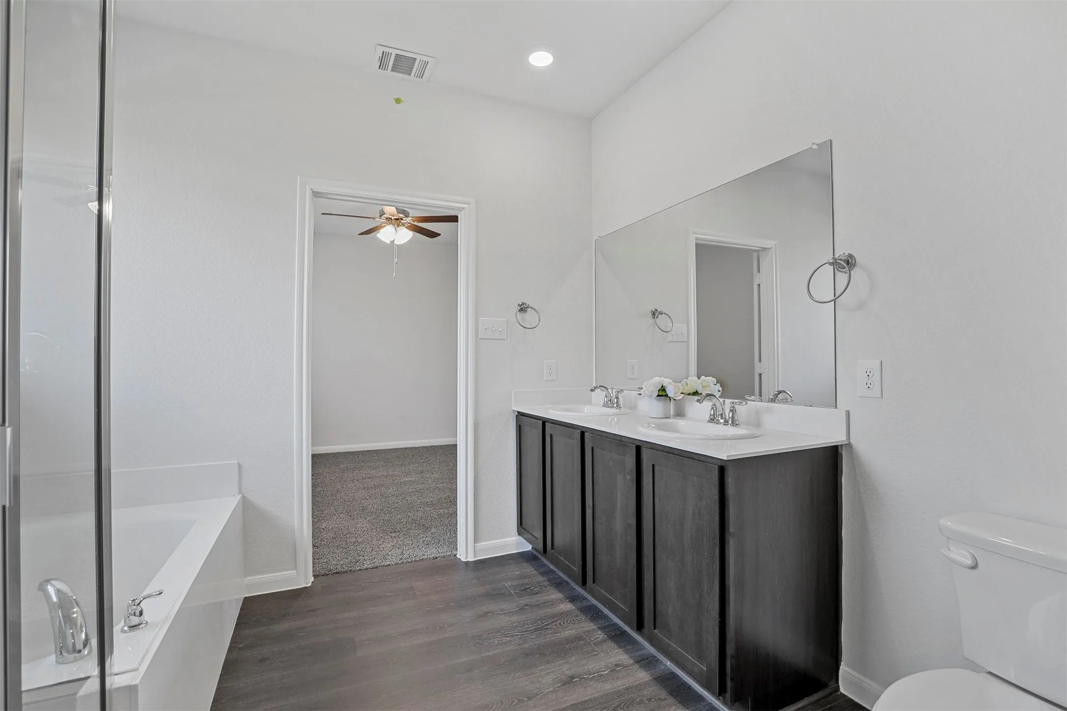 Spa-inspired primary bathroom featuring a double-sink vanity with dark cabinetry and white quartz countertop, large framed mirror with integrated lighting, walk-in shower with glass enclosure, soaking tub, and dark wood-finish flooring throughout