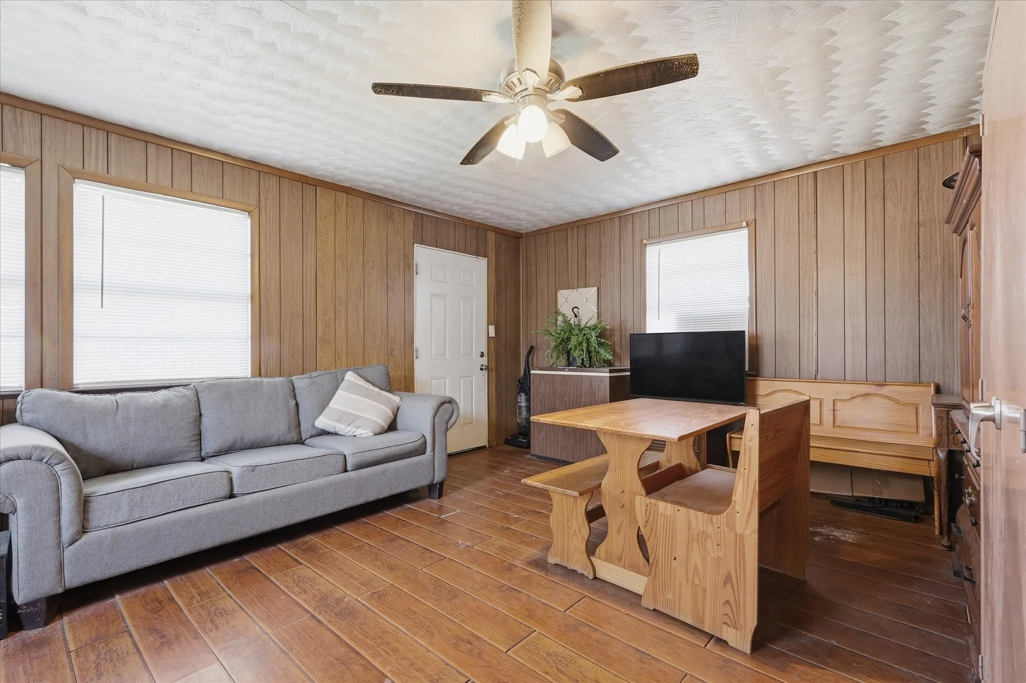 Living area with warm wood-look flooring, paneled walls, ceiling fan, and multiple windows that bring in natural light. The space offers a comfortable everyday layout with room for seating, dining, and media, creating a practical central gathering area with classic character.
