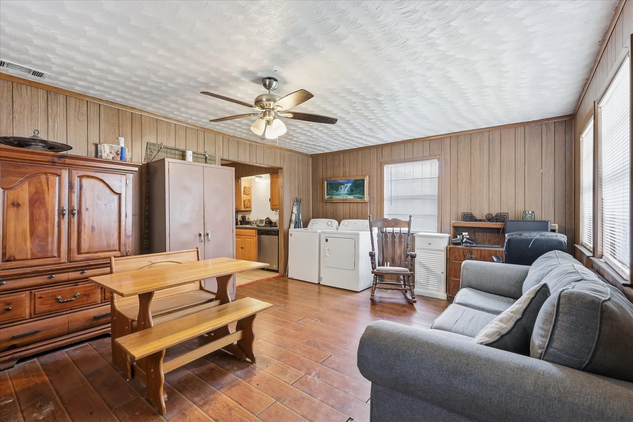 Spacious living and dining area with warm flooring, paneled walls, ceiling fan, and an easy flow toward the kitchen. The room offers flexible use for relaxing, dining, working, or entertaining, with natural light and a comfortable layout that can adapt to a variety of buyer needs.