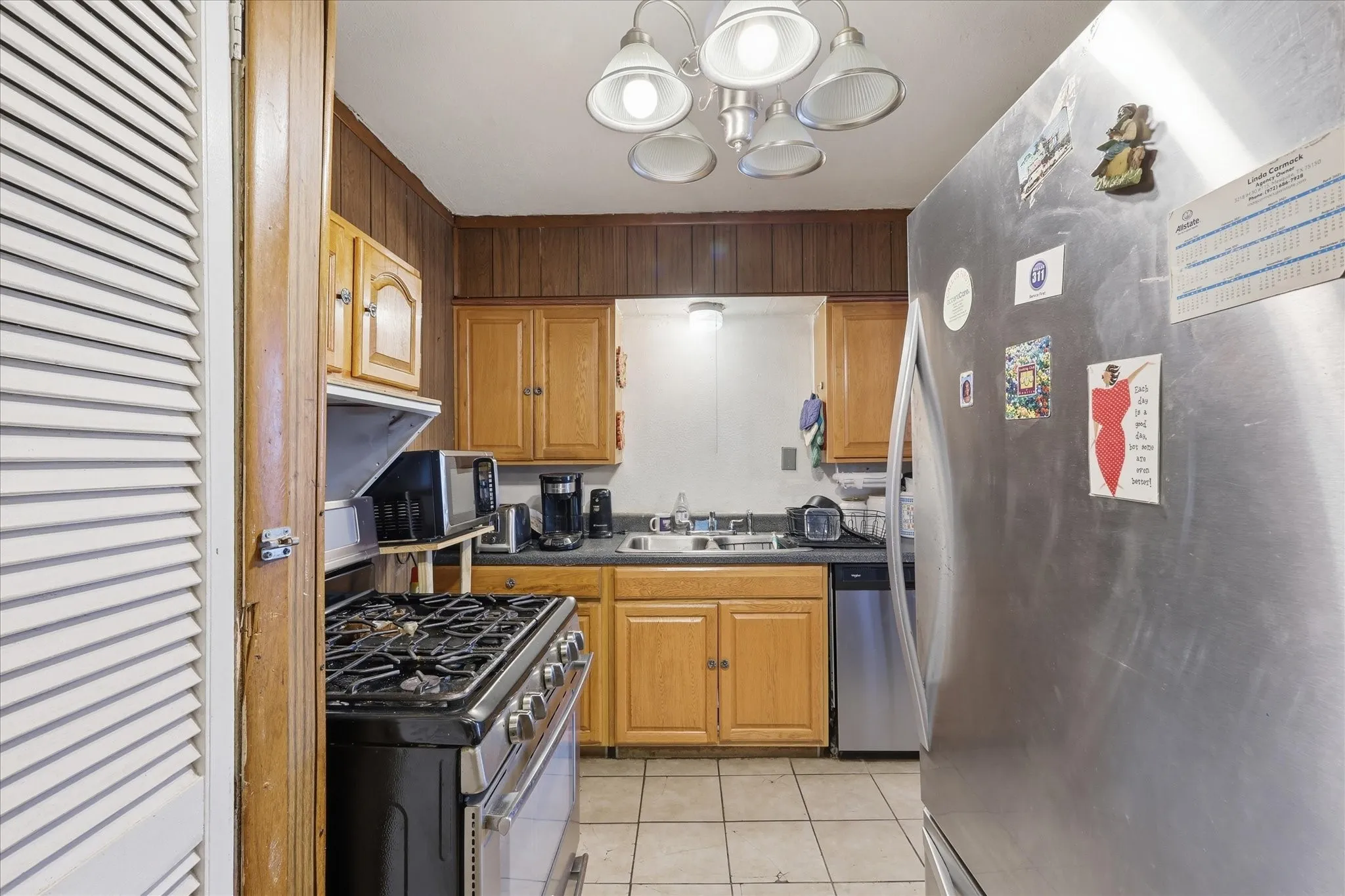 Kitchen view featuring wood cabinetry, tile flooring, stainless-style appliances, sink area, dishwasher, range, and refrigerator space. The compact layout keeps appliances and prep areas within easy reach, creating an efficient workspace for daily cooking. Overhead lighting, cabinet storage, and nearby access to the dining area make the kitchen practical and convenient.