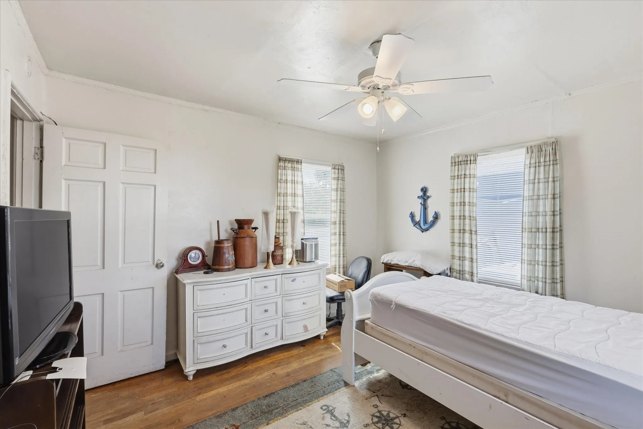 Bedroom view featuring wood flooring, white walls, ceiling fan, closet door, dresser space, and two windows with patterned curtains. The room feels bright and functional, with enough wall space for furniture placement and personal décor. Its practical size and natural light make it well suited for a bedroom, guest room, office, hobby space, or other flexible use.