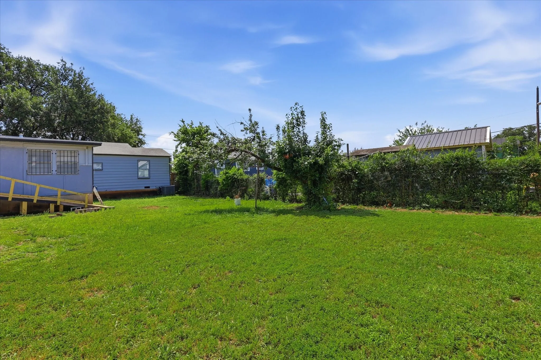Expansive rear yard view with the home positioned to one side and a wide stretch of grass extending across the property. Mature shrubs and trees line the perimeter, creating a natural backdrop and added privacy. This photo showcases the lot’s openness, sunlight, and versatility, making it easy to envision outdoor dining, play space, gardens, or future improvements.