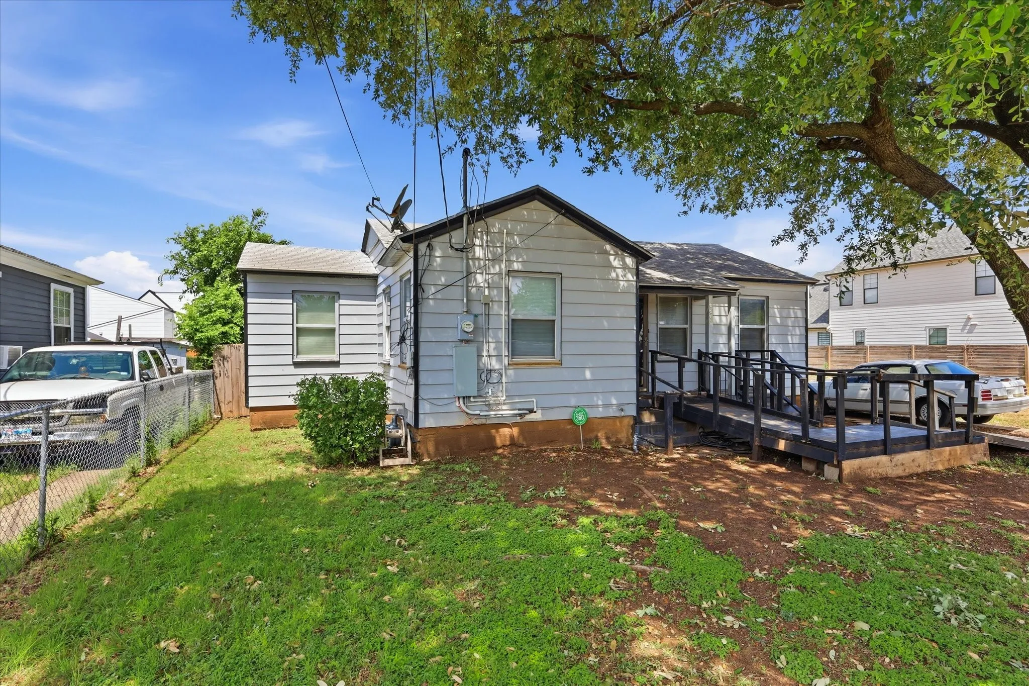 Side/front exterior view showing the home’s traditional structure, shaded porch area, ramped entry, side yard, and neighboring homes. The generous lot layout is visible from this angle, with green lawn space, mature tree coverage, and convenient access around the property. A strong perspective for showing both the home’s footprint and usable outdoor areas.