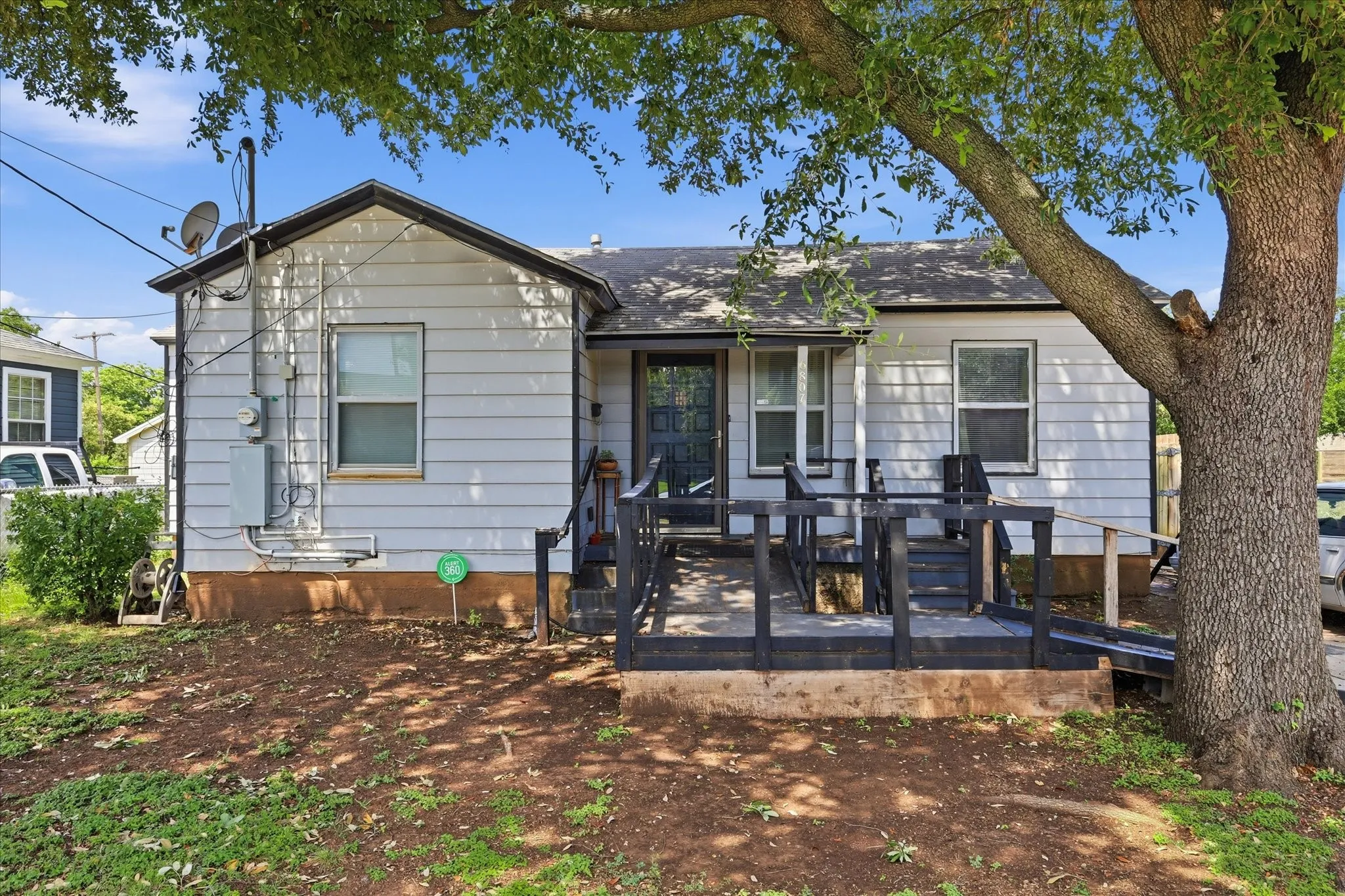 Straight-on front elevation of this traditional Dallas home, featuring white siding, black trim, a covered front entry, and a ramped porch approach beneath a large shade tree. The front yard offers space for landscaping improvements, while the home’s simple classic profile creates an inviting opportunity for buyers seeking character, location, and future potential.
