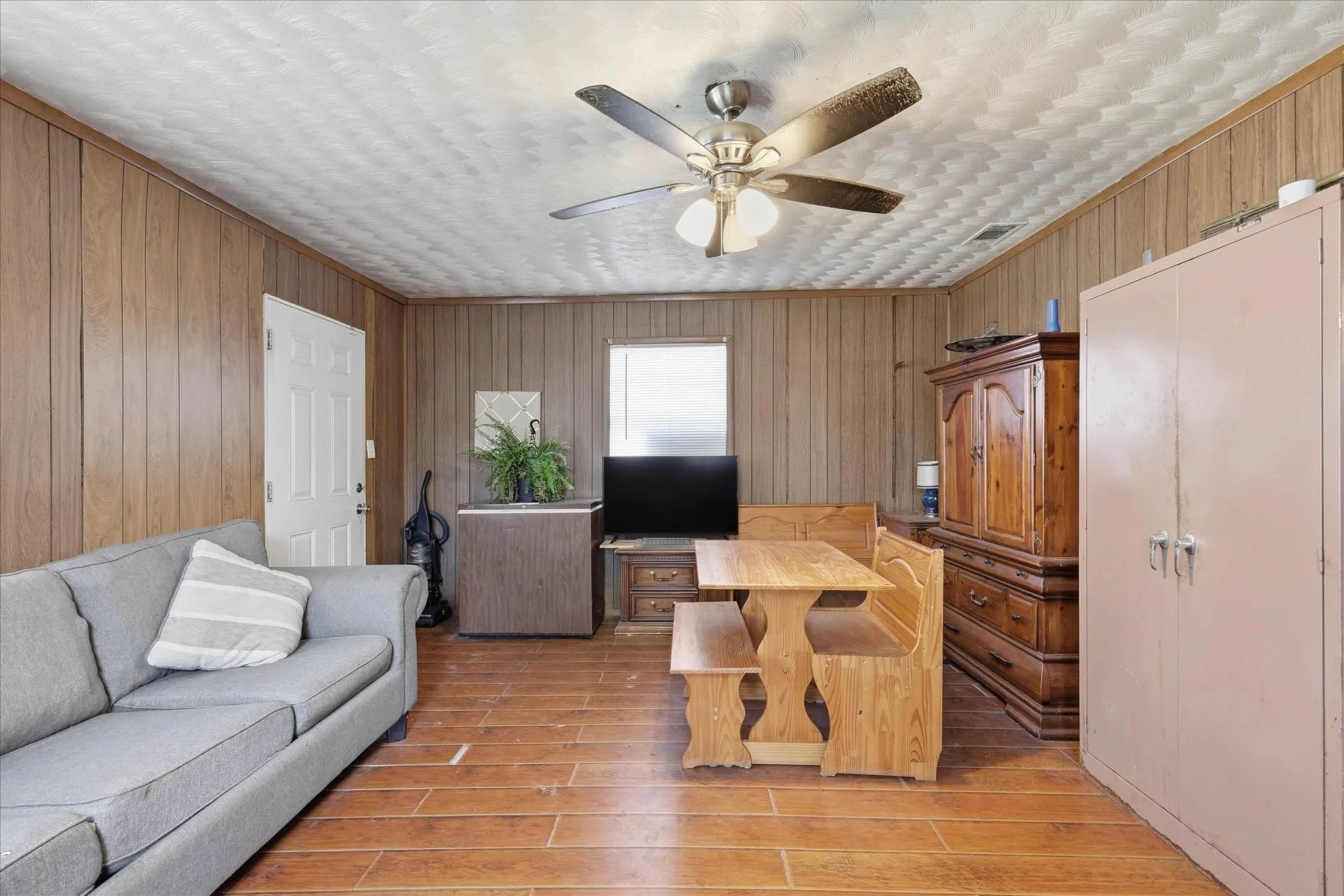 Flexible living and dining area with warm wood-look flooring, paneled walls, ceiling fan, and multiple furniture zones. This room offers a practical central gathering space with room for seating, dining, media, storage, or everyday relaxation. Natural light from the window helps brighten the space, while the layout provides versatility for buyers who want a comfortable living area that can adapt to their needs.