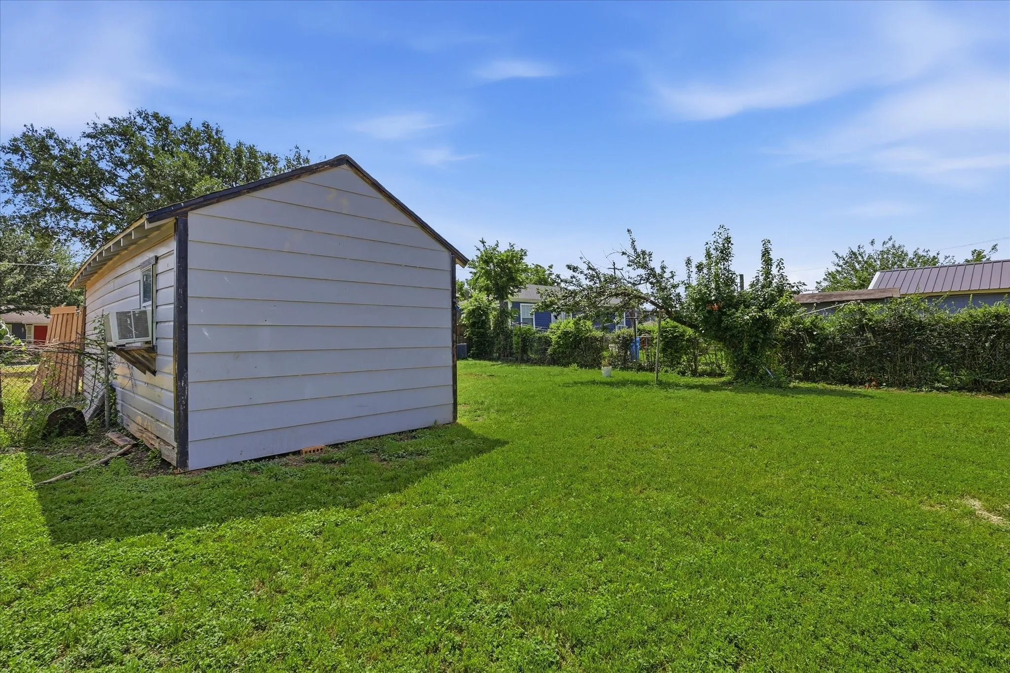 Backyard view from beside the detached garage, showing the large lawn, blue sky, mature greenery, and surrounding fencing. The space feels open and full of potential, ideal for buyers who value outdoor flexibility. Whether used for entertaining, pets, play, gardening, or simply enjoying a private green space, this backyard adds meaningful value.