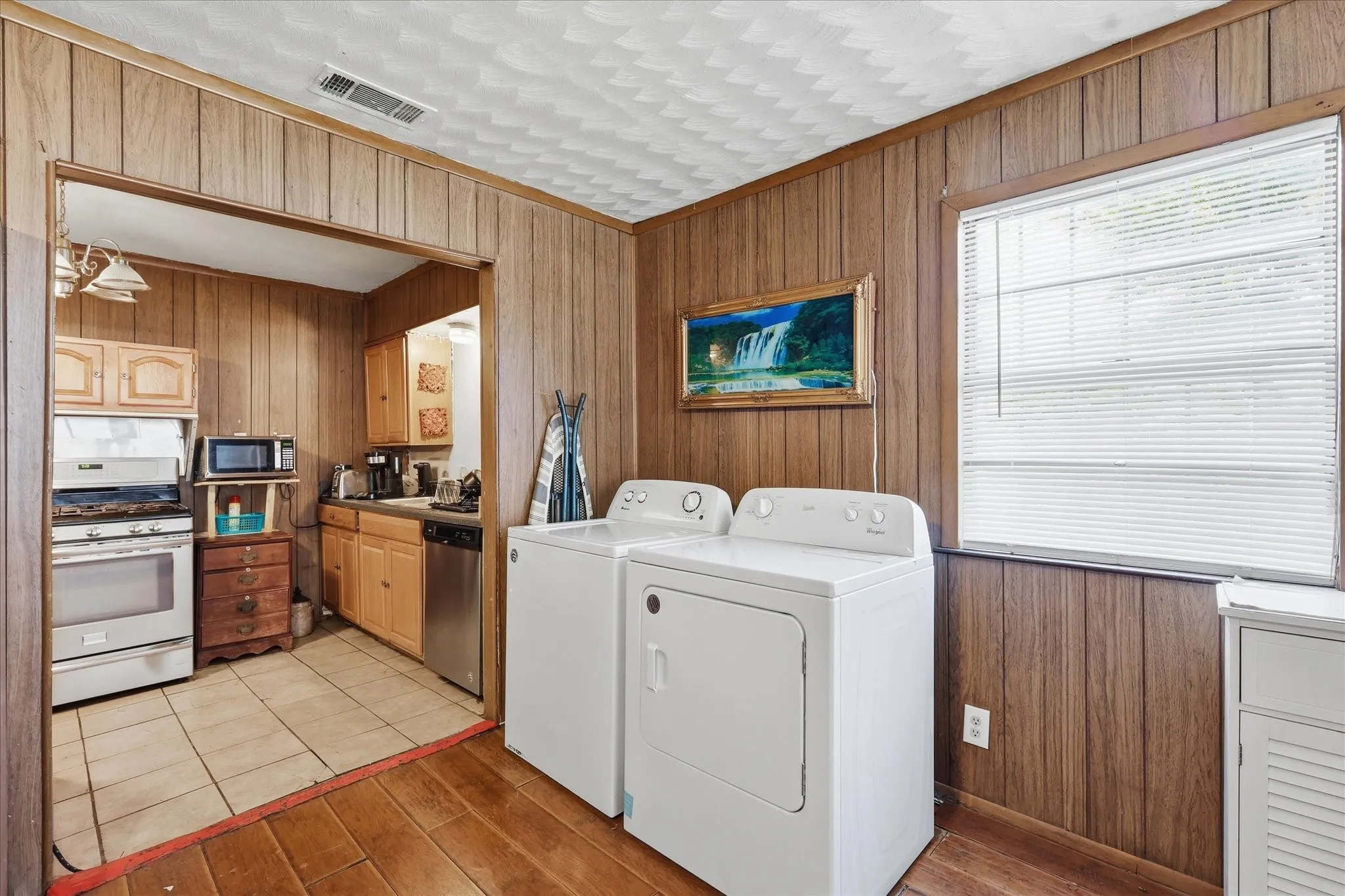 Laundry area with washer and dryer placement, wood-paneled walls, warm wood-look flooring, and convenient access to the kitchen. This space adds valuable everyday function to the home, keeping laundry easily accessible while remaining near the main living and kitchen areas. A window brings in natural light, and the open connection to the kitchen helps the layout feel practical and efficient.