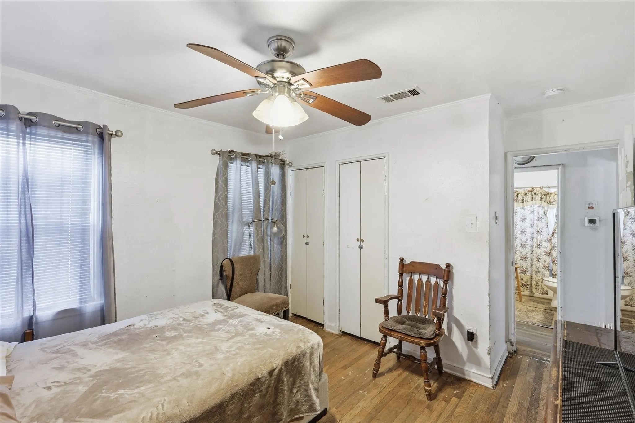 Bedroom view highlighting the ceiling fan, wood flooring, closet doors, multiple windows, and direct sightline toward the bathroom. The room provides functional storage, natural light, and enough space for bedroom furniture plus a small seating area. With white walls and warm floors, this space offers a clean, flexible canvas ready for everyday use or future personal updates.
