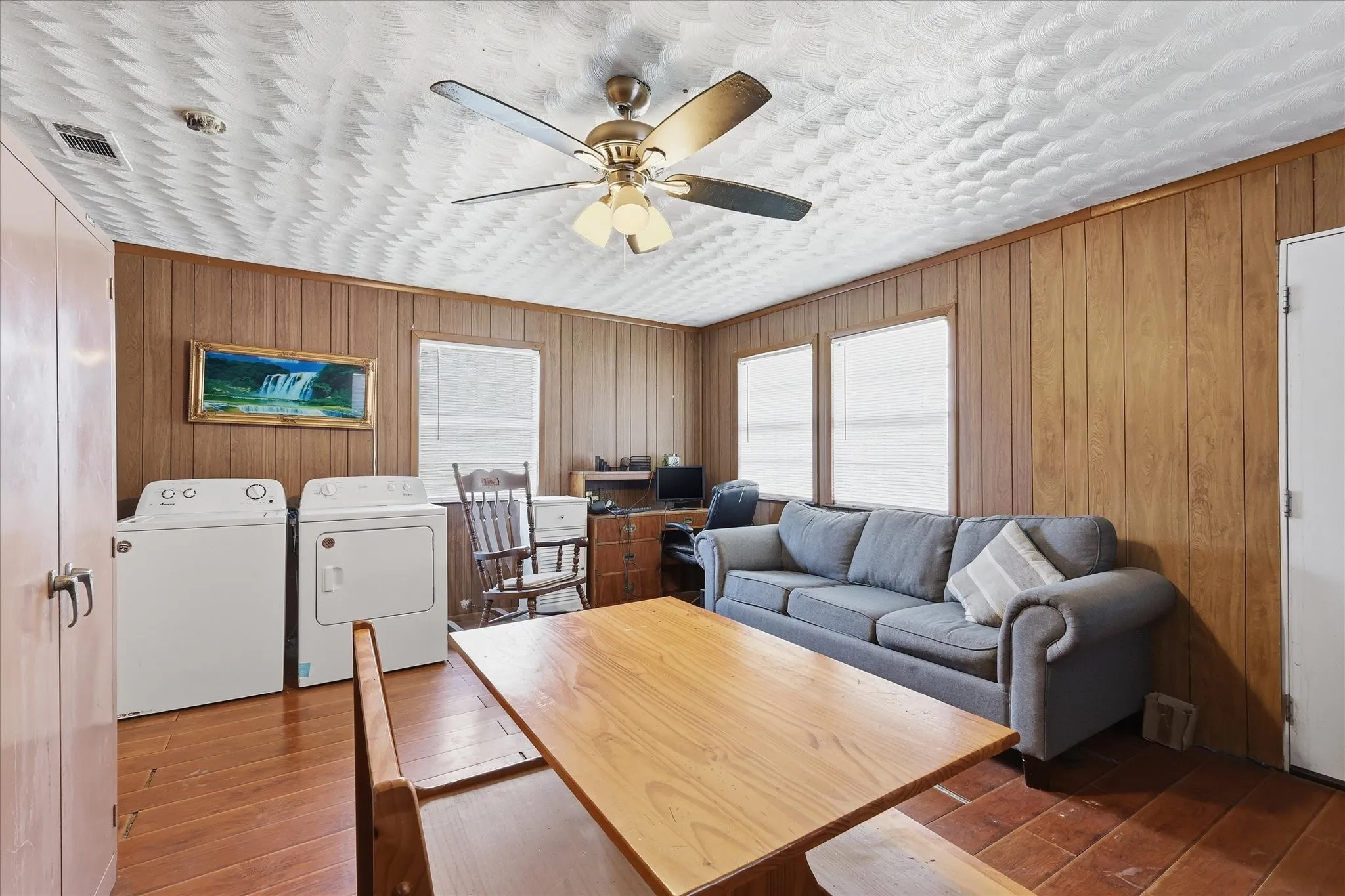 Living room view highlighting the ceiling fan, textured ceiling detail, wood-look floors, paneled walls, and flexible furniture arrangement. With natural light from multiple windows and space for seating, laundry, and work-from-home use, this room offers practical everyday versatility.
