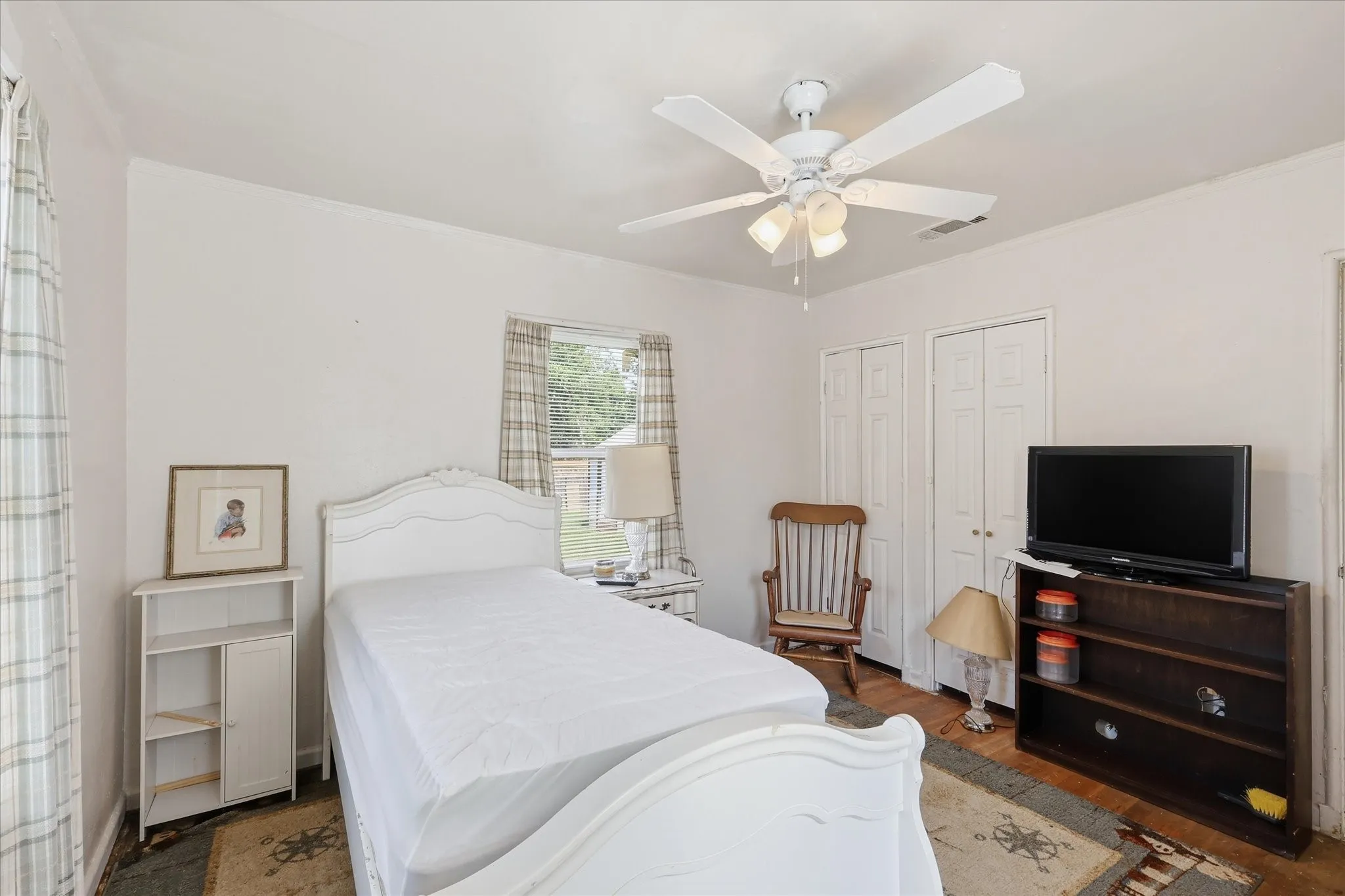 Comfortable bedroom with white walls, ceiling fan, closet doors, wood flooring, and natural light from the window. The room includes space for a bed, side table, seating, dresser, and media furniture, offering an easy-to-use layout with multiple furnishing options. Neutral finishes help create a simple, bright backdrop ready for a buyer’s personal style.
