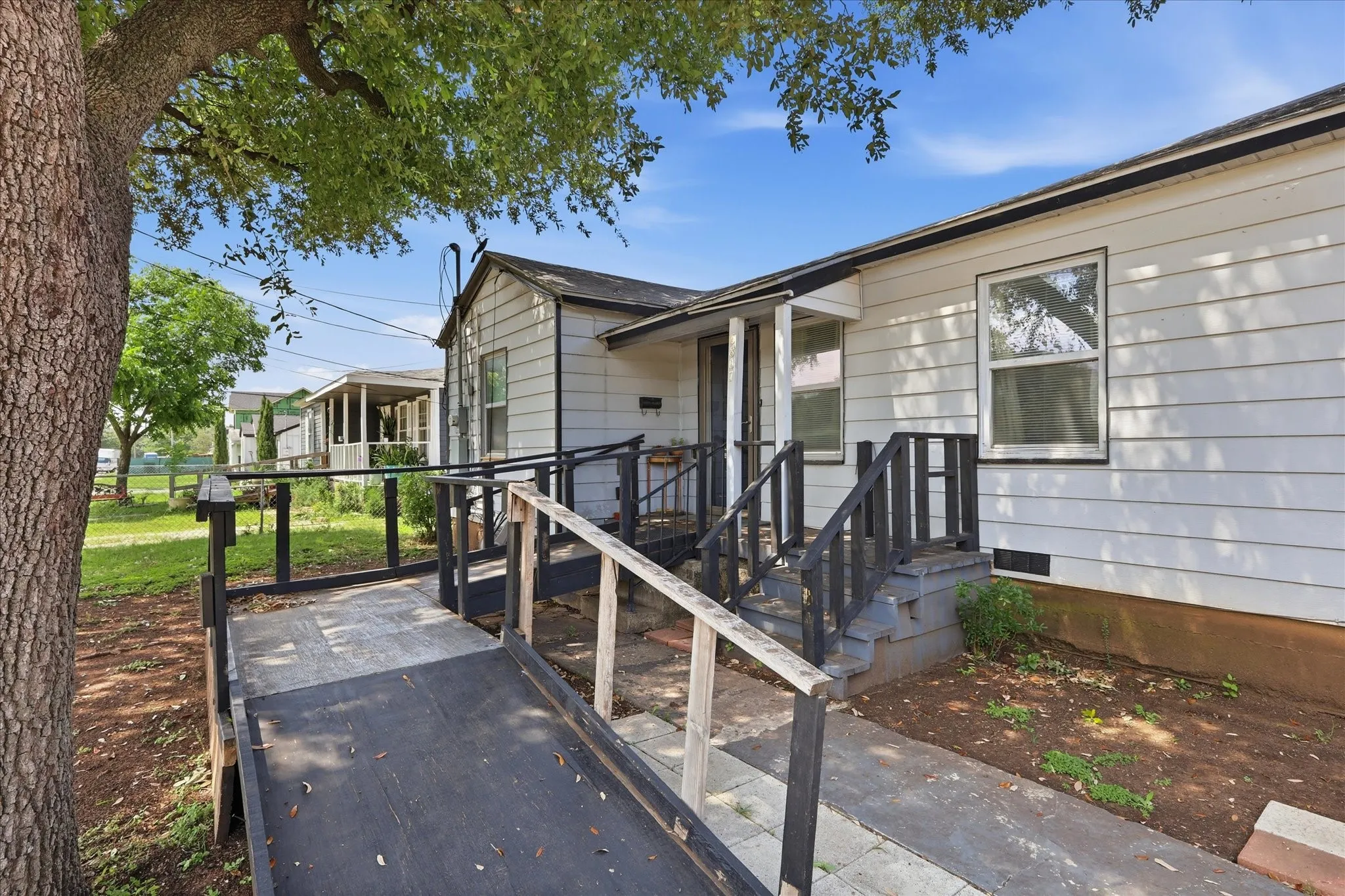 A welcoming first look at 6807 Prosper Street, showcasing the home’s classic exterior, mature shade tree, and convenient ramped approach to the covered front entry. The black railing, porch area, and wide walkway create a practical arrival experience, while the established neighboring homes and leafy setting give the property a comfortable Dallas neighborhood feel.