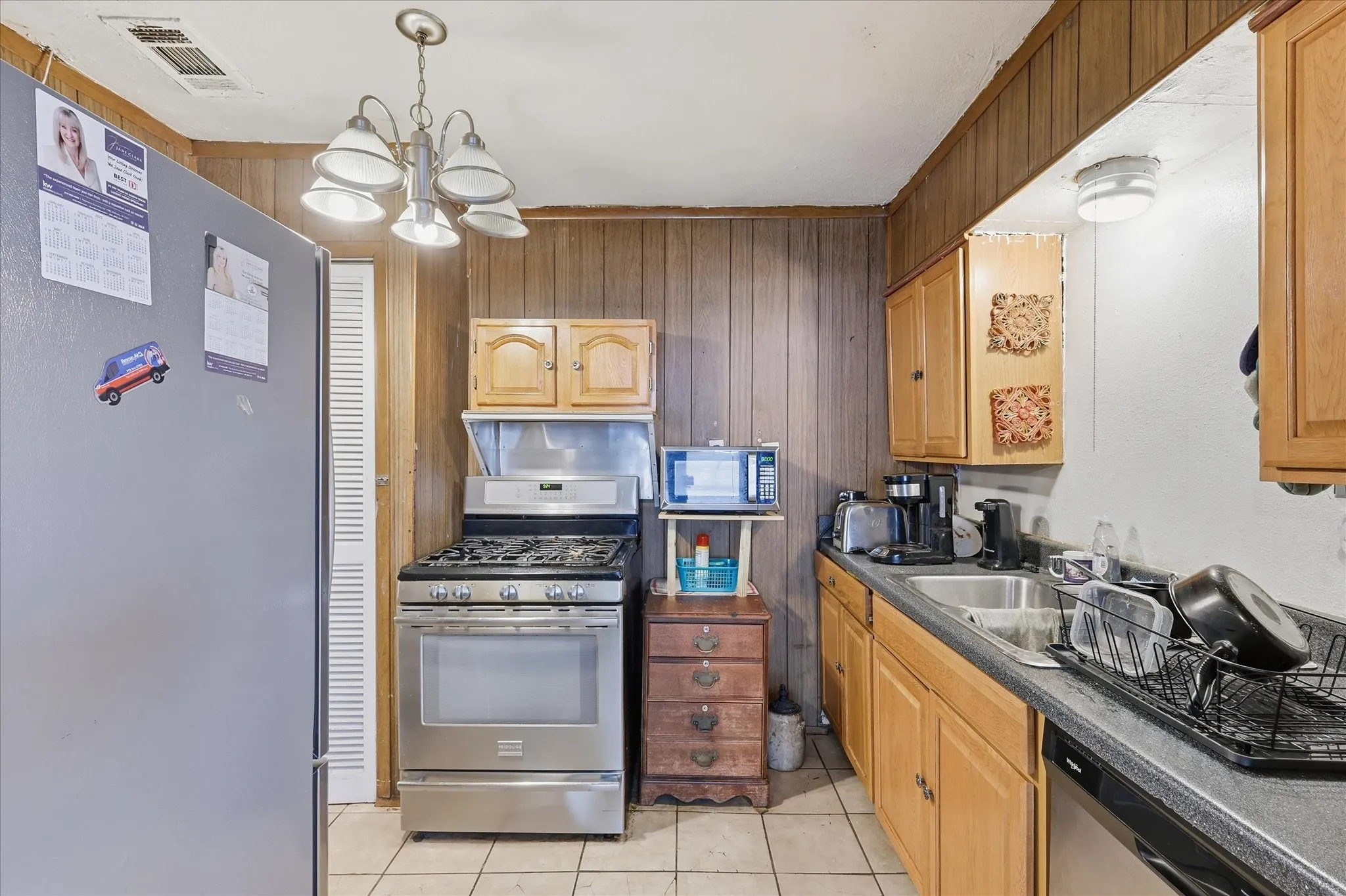 Functional kitchen with tile flooring, wood cabinetry, stainless-style range, dishwasher, sink, refrigerator, and countertop workspace. The layout offers practical storage and efficient flow for meal prep, cleanup, and everyday cooking. Warm cabinet tones and paneled accents add classic character, while the adjacent dining area provides convenient access for serving and gathering.