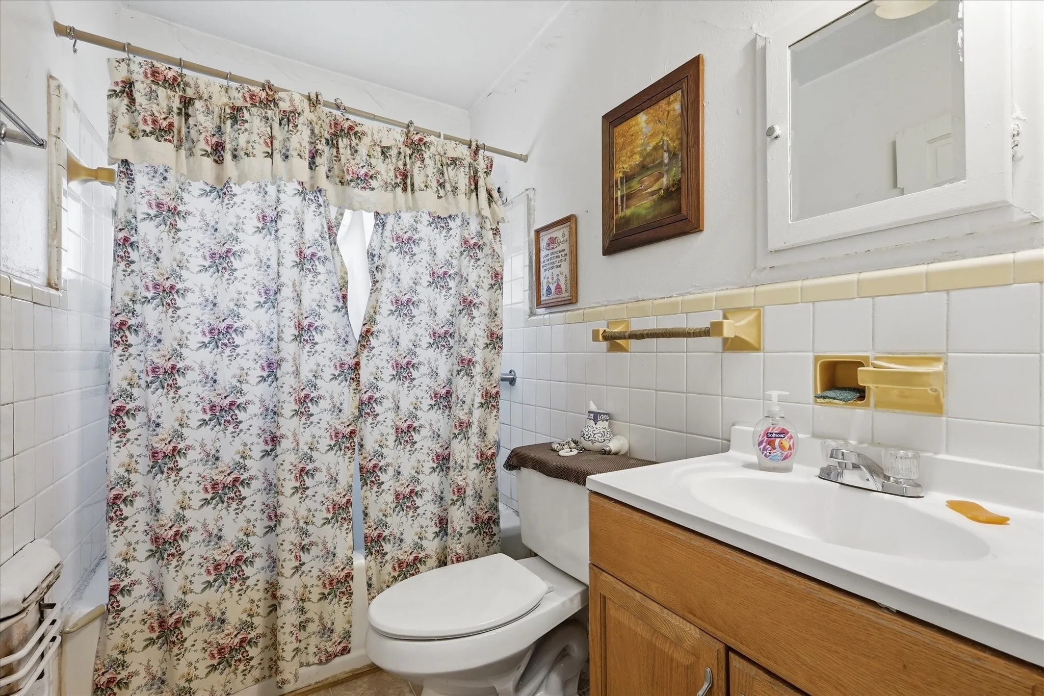 Full bathroom with a tub/shower combination, tile surround, floral shower curtain, vanity storage, and a bright white sink area. The room offers a practical layout with essential daily function, including mirror, towel bars, built-in wall accents, and neutral tile details. Its compact footprint keeps everything within easy reach while providing the convenience of a full bath for the home.