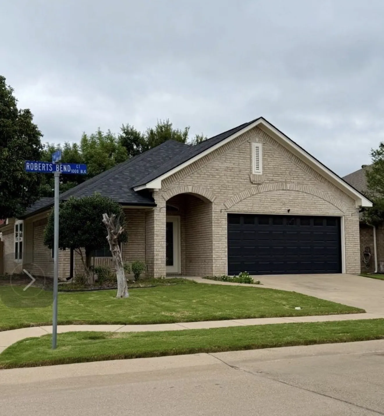 Cream brick exterior, arched entryway, dark asphalt shingle roof, two-car garage, and a concrete driveway