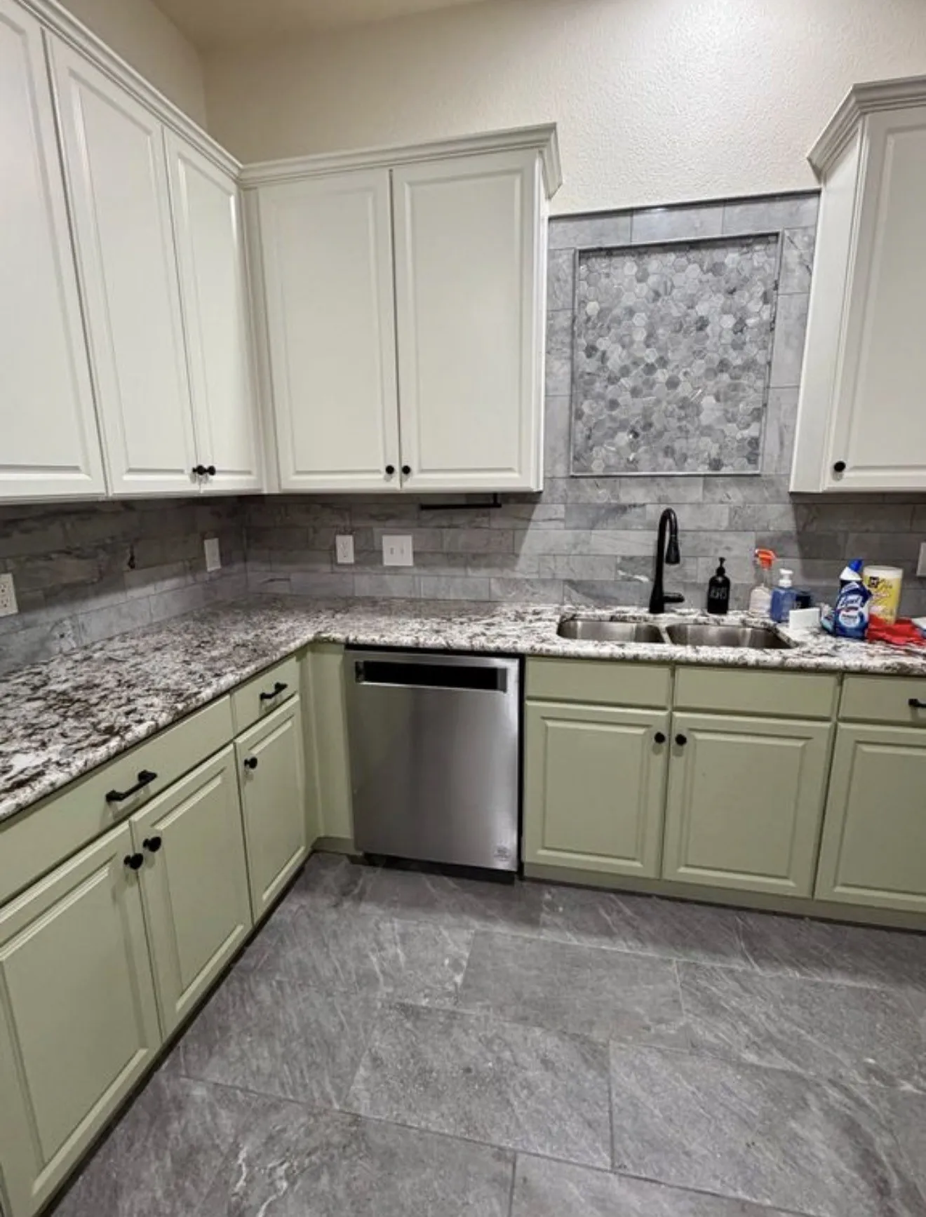 White shaker upper cabinetry, sage lower cabinetry, granite countertops, tile backsplash with decorative mosaic inlay, and stainless steel dishwasher