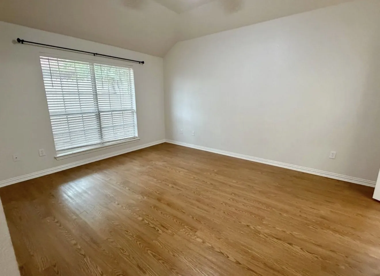 Vaulted ceiling, wood-finish flooring, white walls, large window with blinds, and a curtain rod