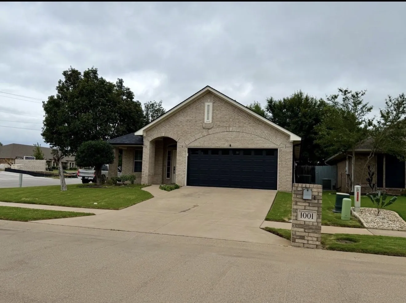 Brick facade home featuring a prominent arched entry, dark garage door, stone mailbox pillar, and mature landscaping