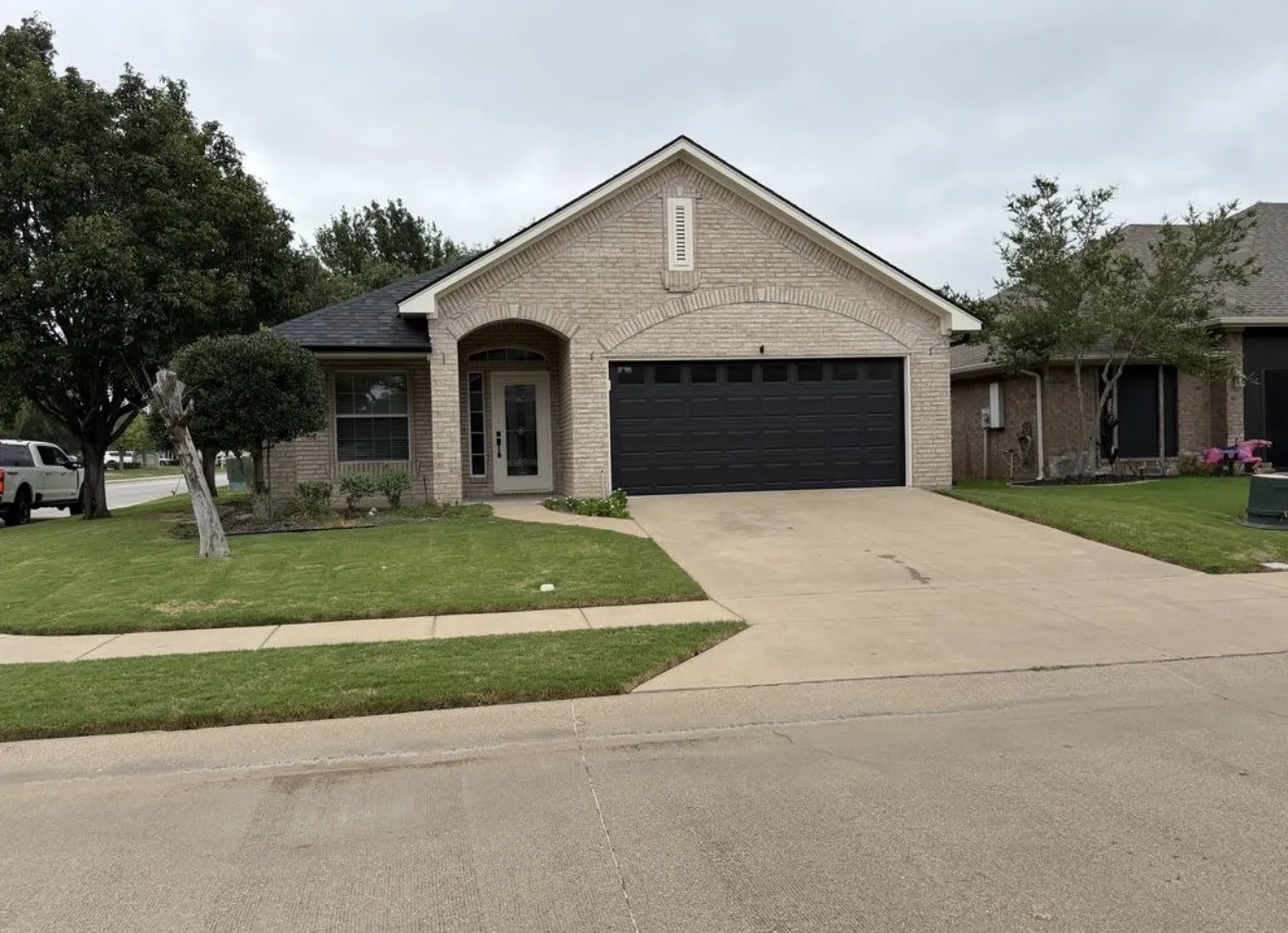 Light-colored brick exterior with an arched entryway, attached two-car garage, concrete driveway, and established front landscaping