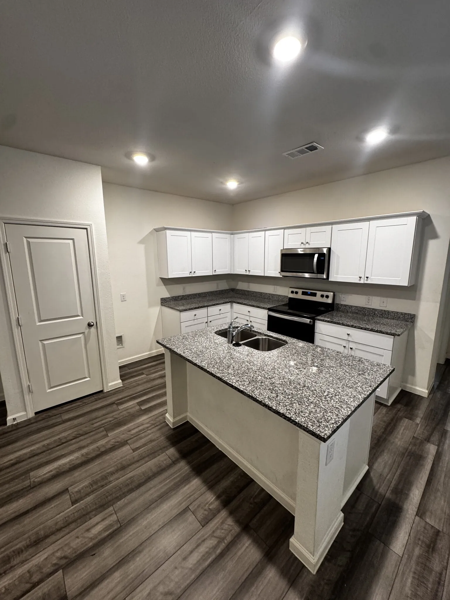 Modern kitchen featuring an island with a stainless steel sink, granite-style countertops, white cabinetry, and wood-finish flooring