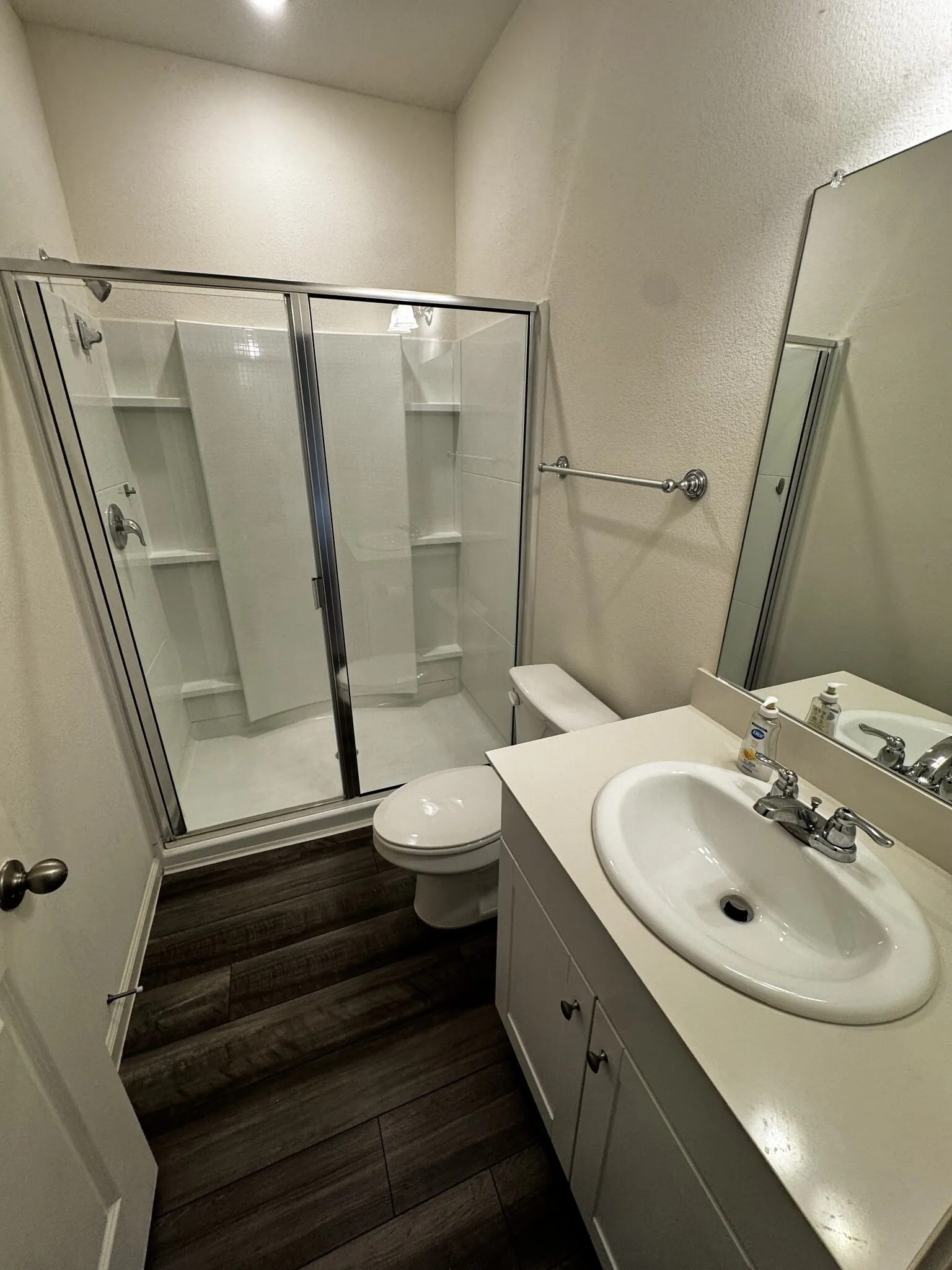 Walk-in shower with glass enclosure, single vanity with integrated sink, wood-finish flooring, and wall-mounted mirror