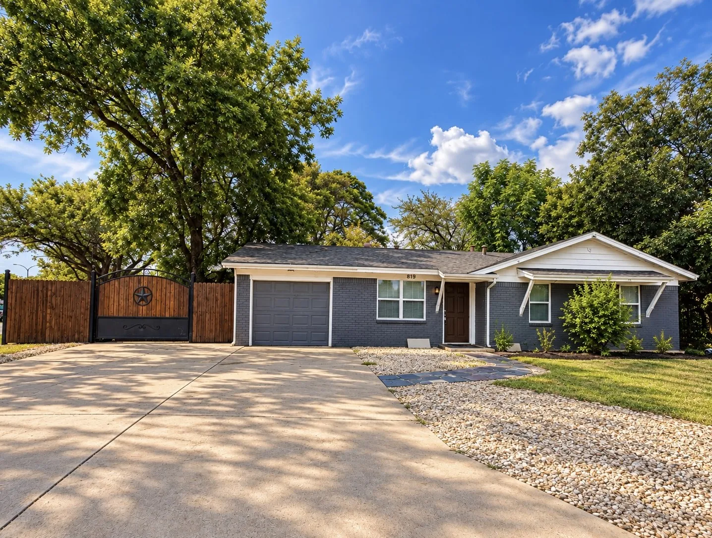 Single-story residence featuring a dark gray painted brick exterior, a driveway with an adjacent gravel path, an attached garage, a wood privacy fence with a decorative gate, and mature trees
