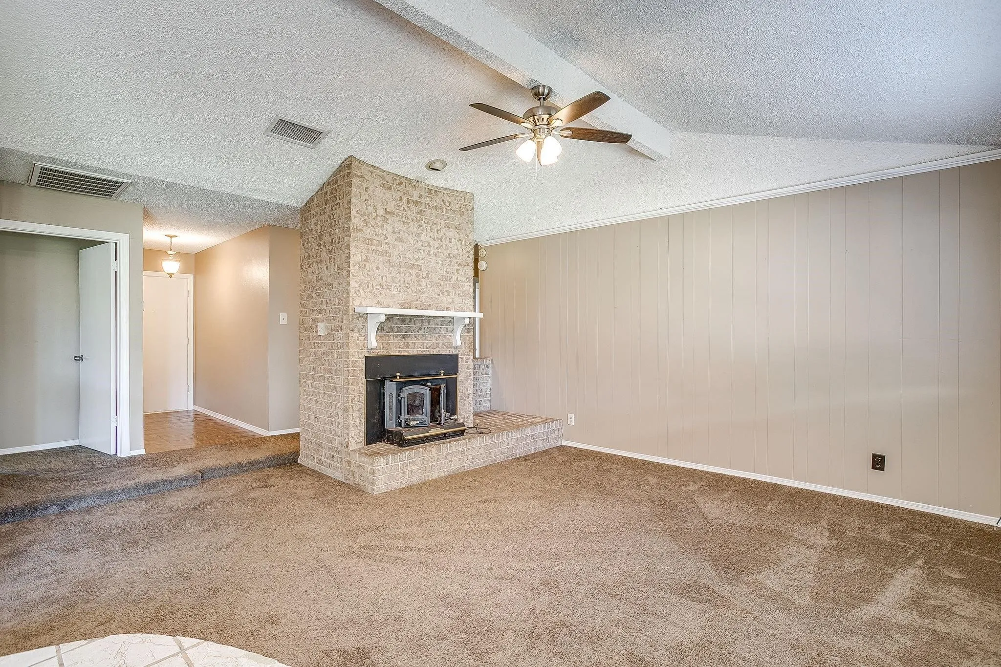 Vaulted ceiling with an exposed beam, brick fireplace with a wood-burning insert, and a ceiling fan with integrated lighting