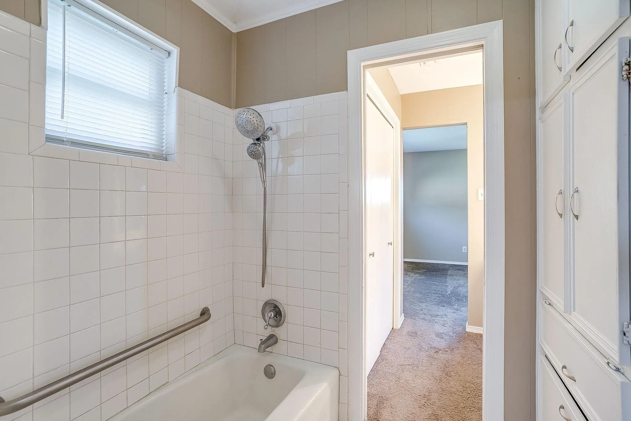 Bathtub with white tile surround, window with horizontal blinds, grab bar, and built-in storage cabinetry