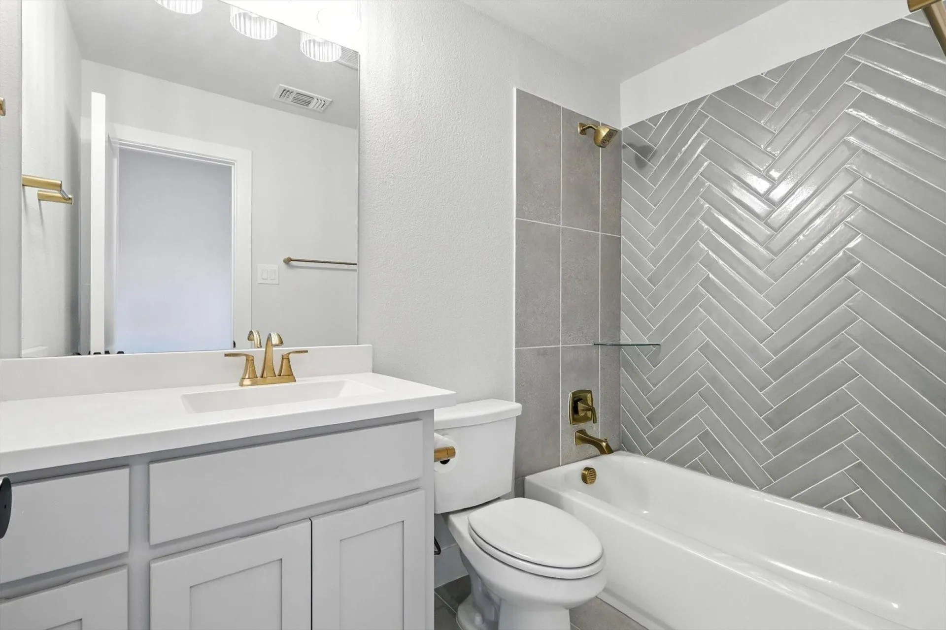 Herringbone-patterned tile shower surround, integrated bathtub, white vanity with gold-tone hardware, white countertop with integral sink, and contemporary lighting fixture