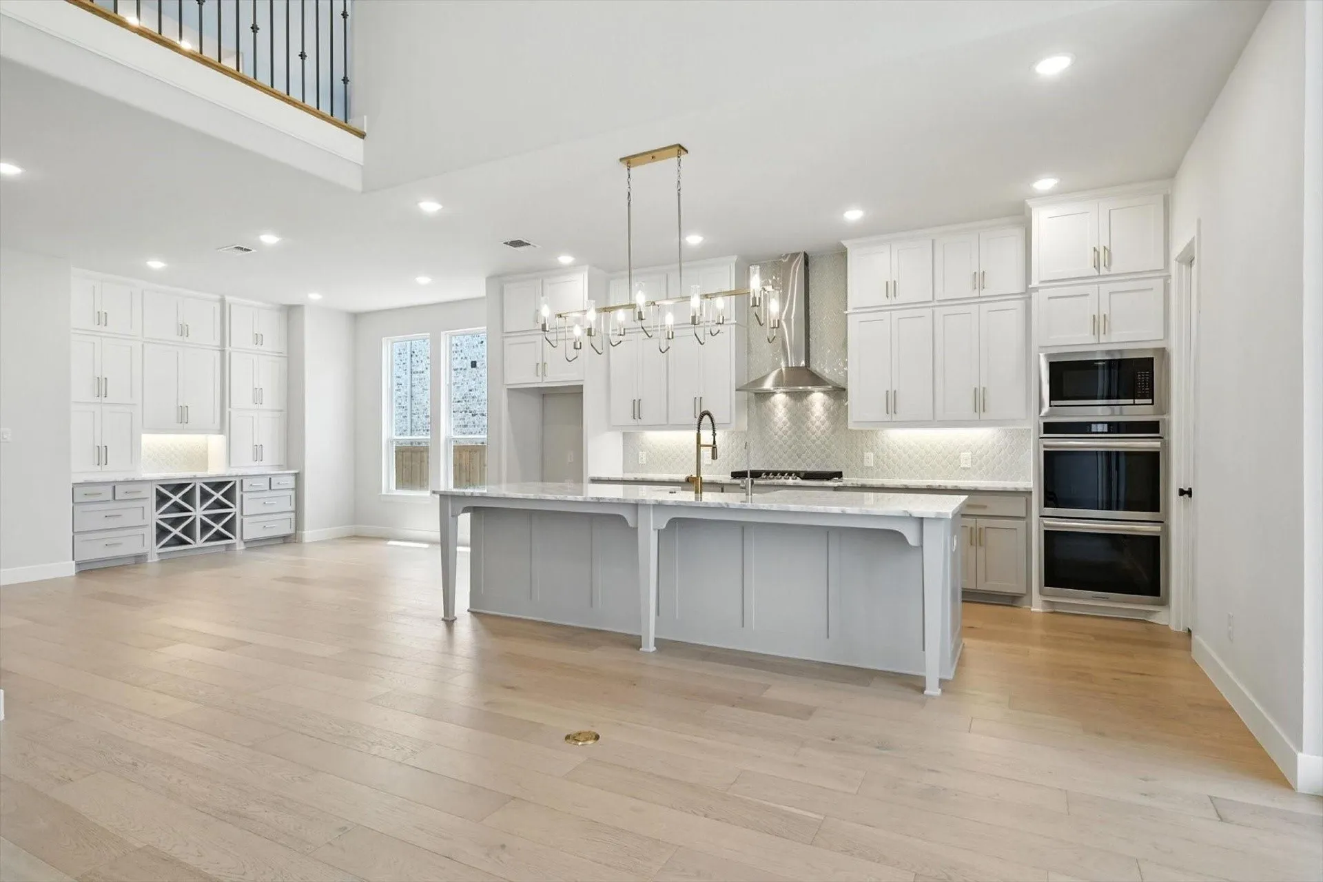 Expansive open-concept space featuring light wood-finish flooring, a kitchen island with a stone countertop, white cabinetry with modern hardware, and stainless steel built-in appliances
