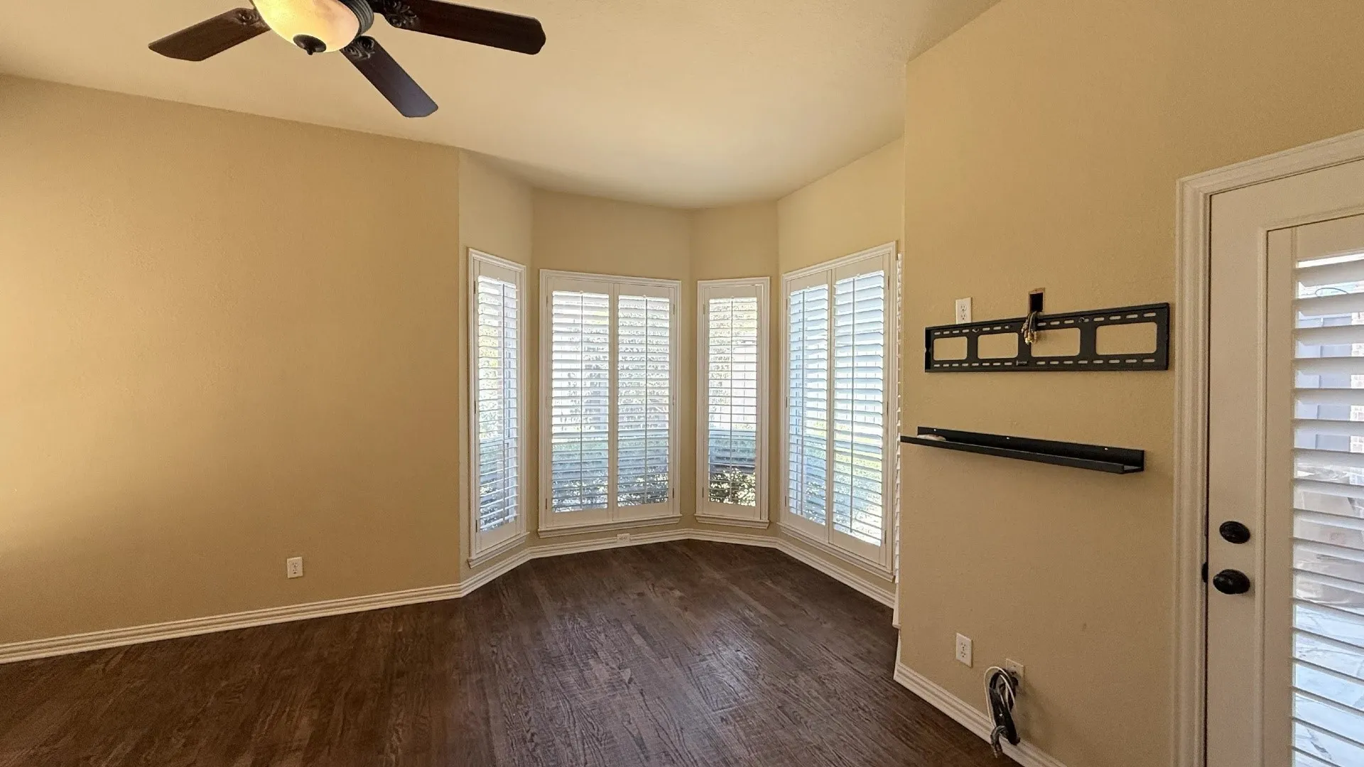 Bay window with plantation shutters, wood-finish flooring, ceiling fan with integrated lighting, and a rear door with integrated blinds