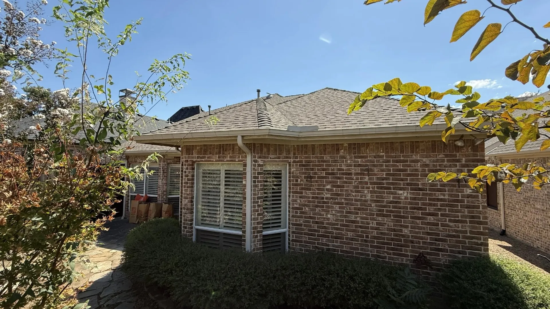 Brick exterior, multiple windows with louvered blinds, shingled roof, rain gutters, and established landscaping