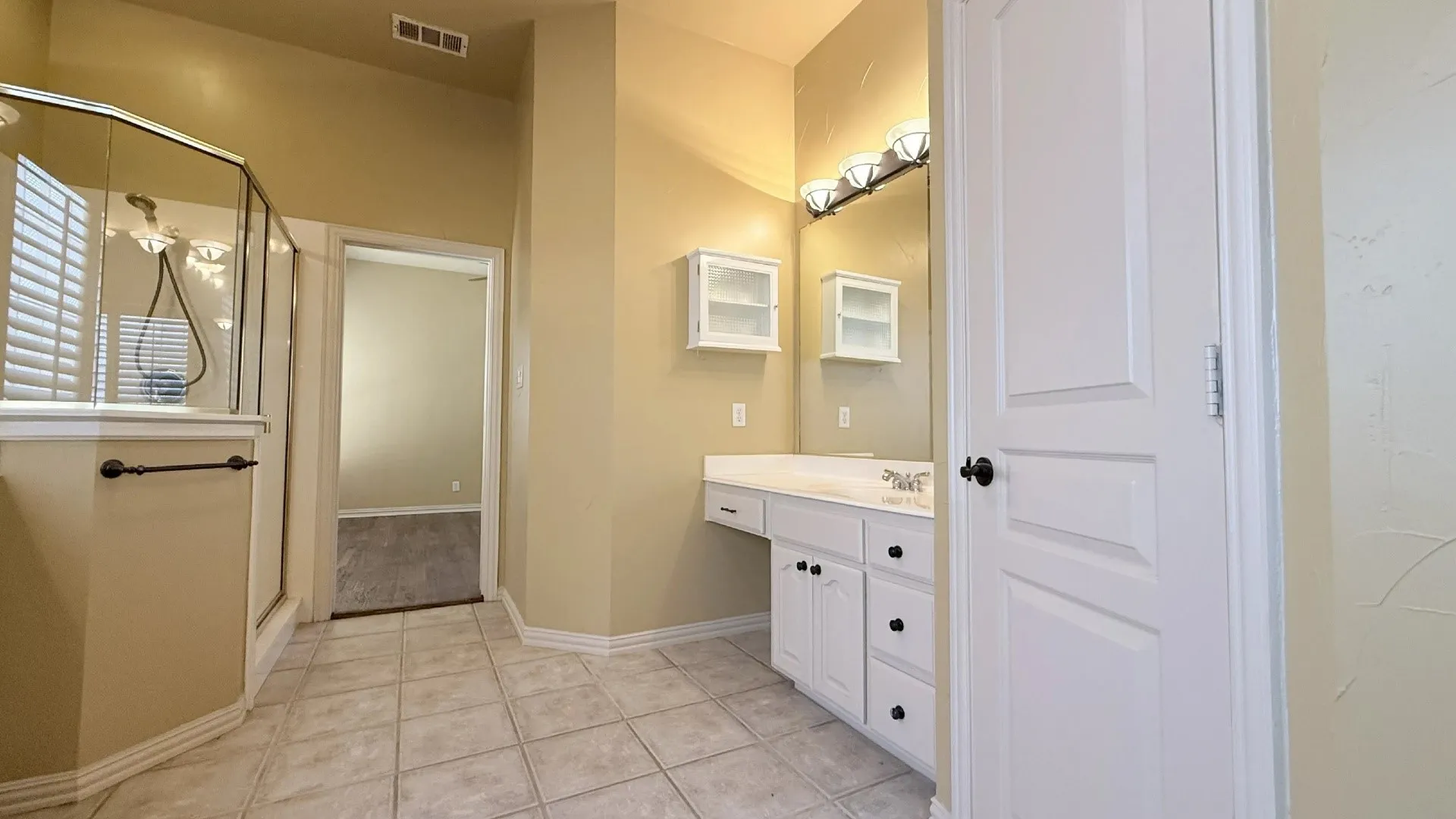 Tiled flooring, glass-enclosed shower, white vanity with integrated sink, wall-mounted mirror, and white cabinetry