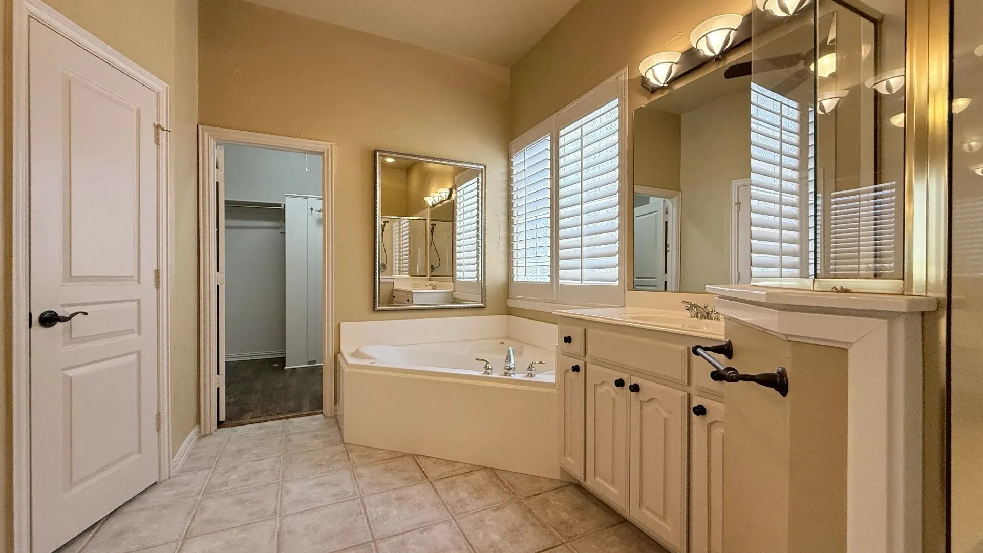Spacious bathroom featuring a corner soaking tub, dual vanity with cabinet storage, large mirror, and window with plantation shutters