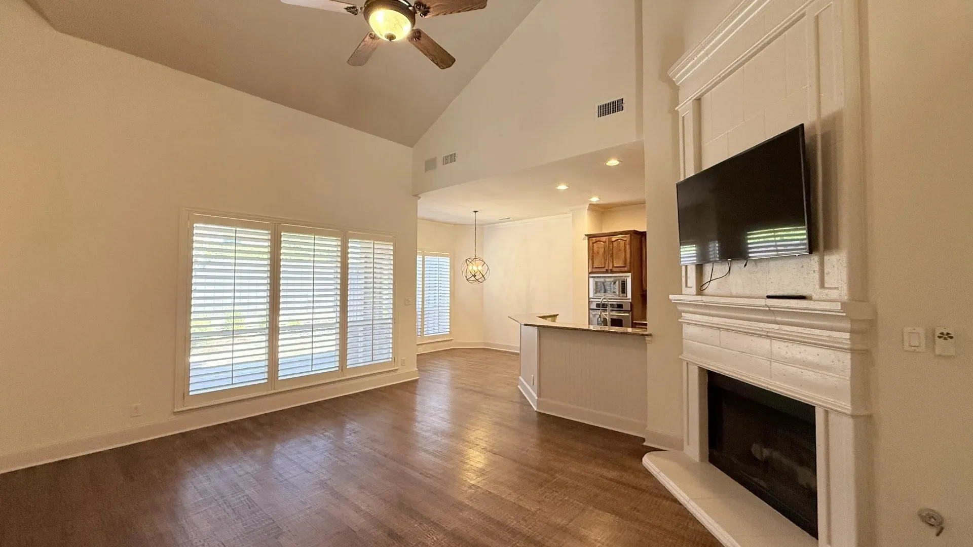 Vaulted ceiling with a ceiling fan, wood-finish flooring, fireplace with a decorative mantel, multiple windows with plantation shutters, and a built-in kitchen island