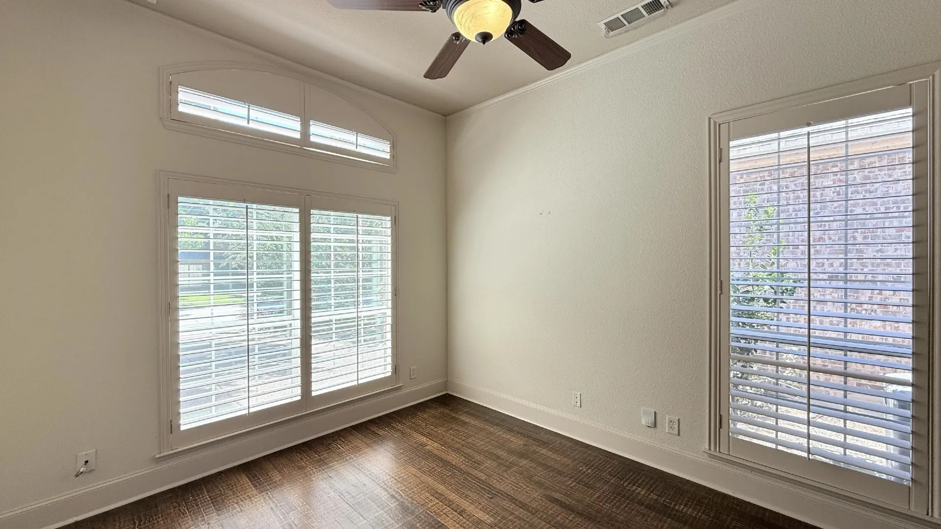Arched window with plantation shutters, additional window with plantation shutters, wood-finish flooring, and a ceiling fan with light fixture