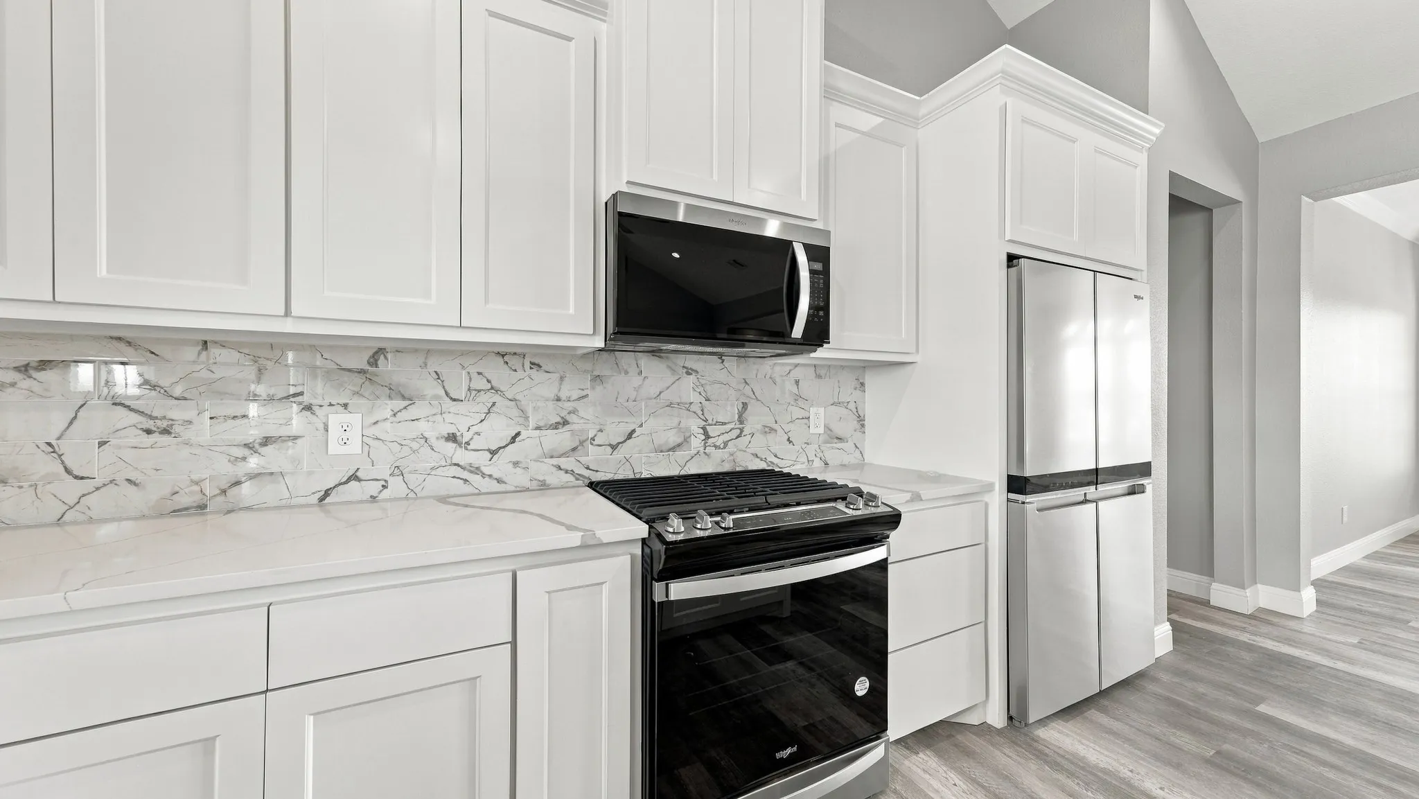 Kitchen featuring white shaker-style cabinetry, a stone-patterned tile backsplash, quartz countertops, stainless steel appliances, and wood-finish flooring under a vaulted ceiling