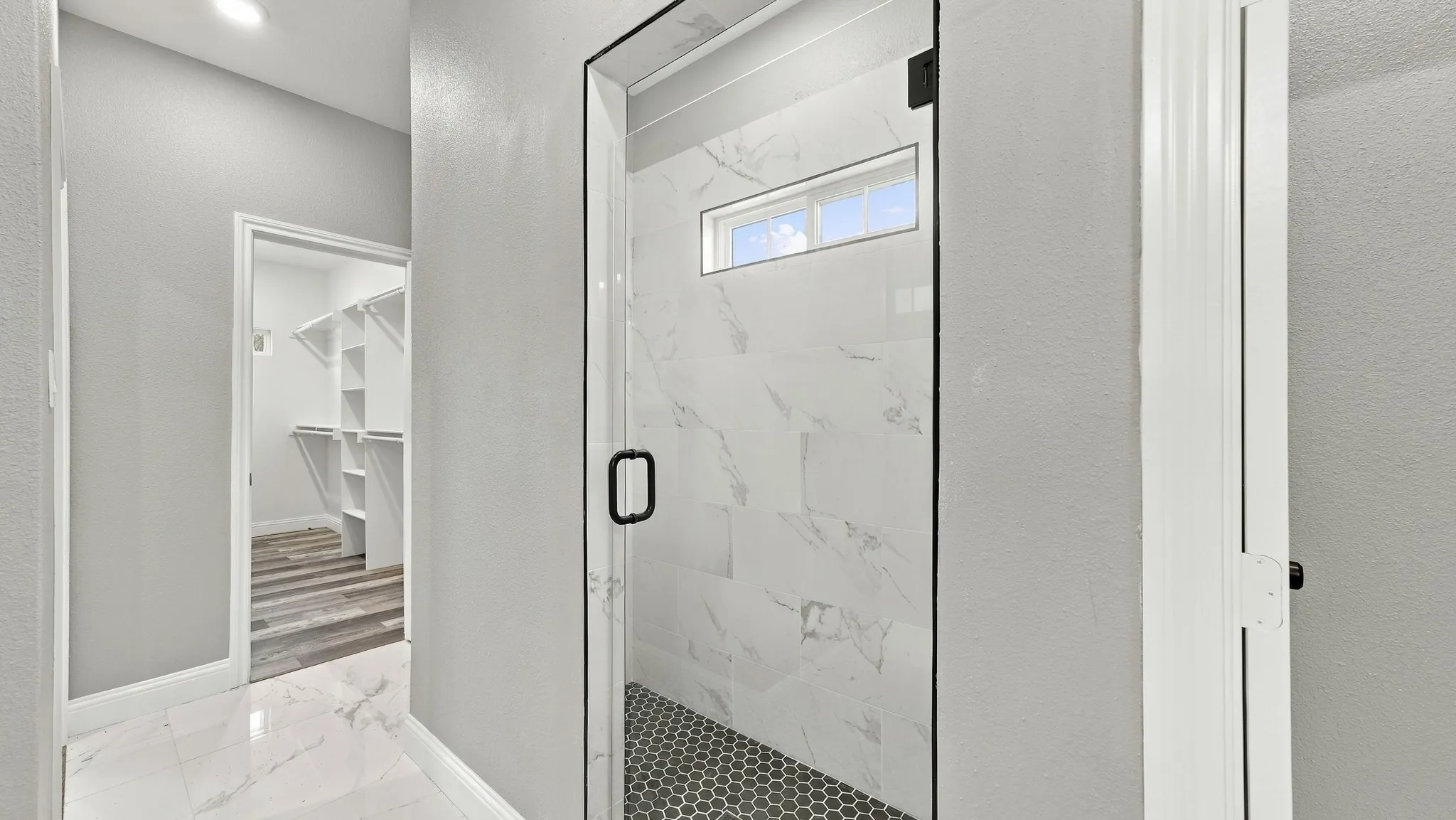 Walk-in shower featuring white tile surround with grey veining, frameless glass door, matte black hardware, and dark hexagonal floor tile