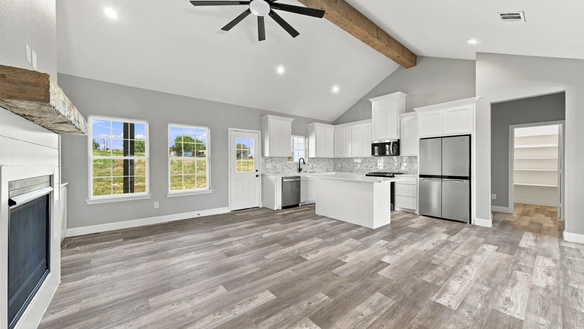 Vaulted ceiling featuring a decorative wood-finish beam, wood-finish flooring, white shaker-style cabinetry, stainless steel appliances, and a fireplace with a stone-finish mantel