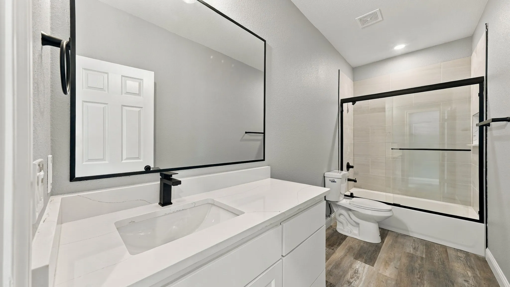 White vanity with a solid-surface countertop and integrated sink, matte black faucet and matching framed mirror, and wood-finish flooring
