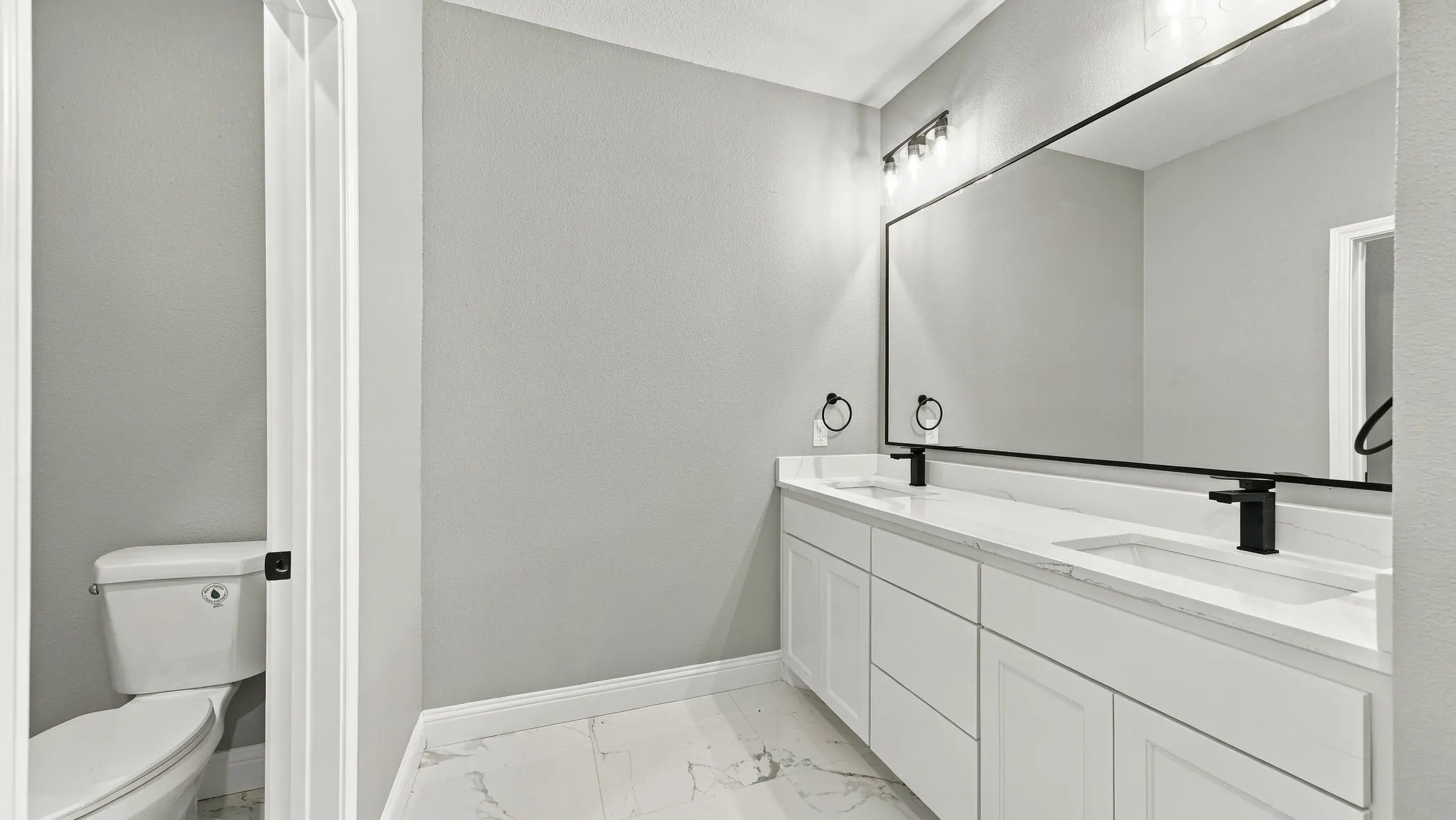 Double vanity with white cabinetry, stone-finish countertops, dual rectangular undermount sinks, matte black fixtures, and a full-width framed mirror