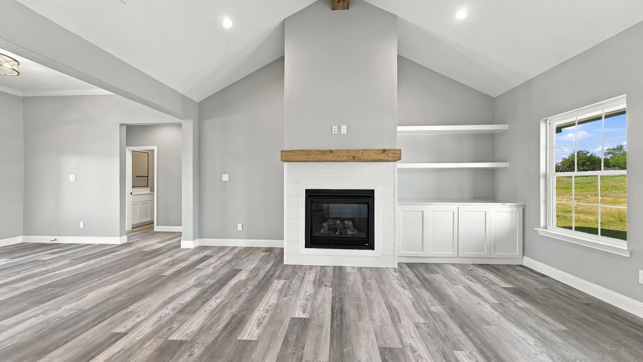 Vaulted ceiling, wood-finish flooring, fireplace with a rustic timber mantel, shiplap surround, and built-in cabinetry with open shelving