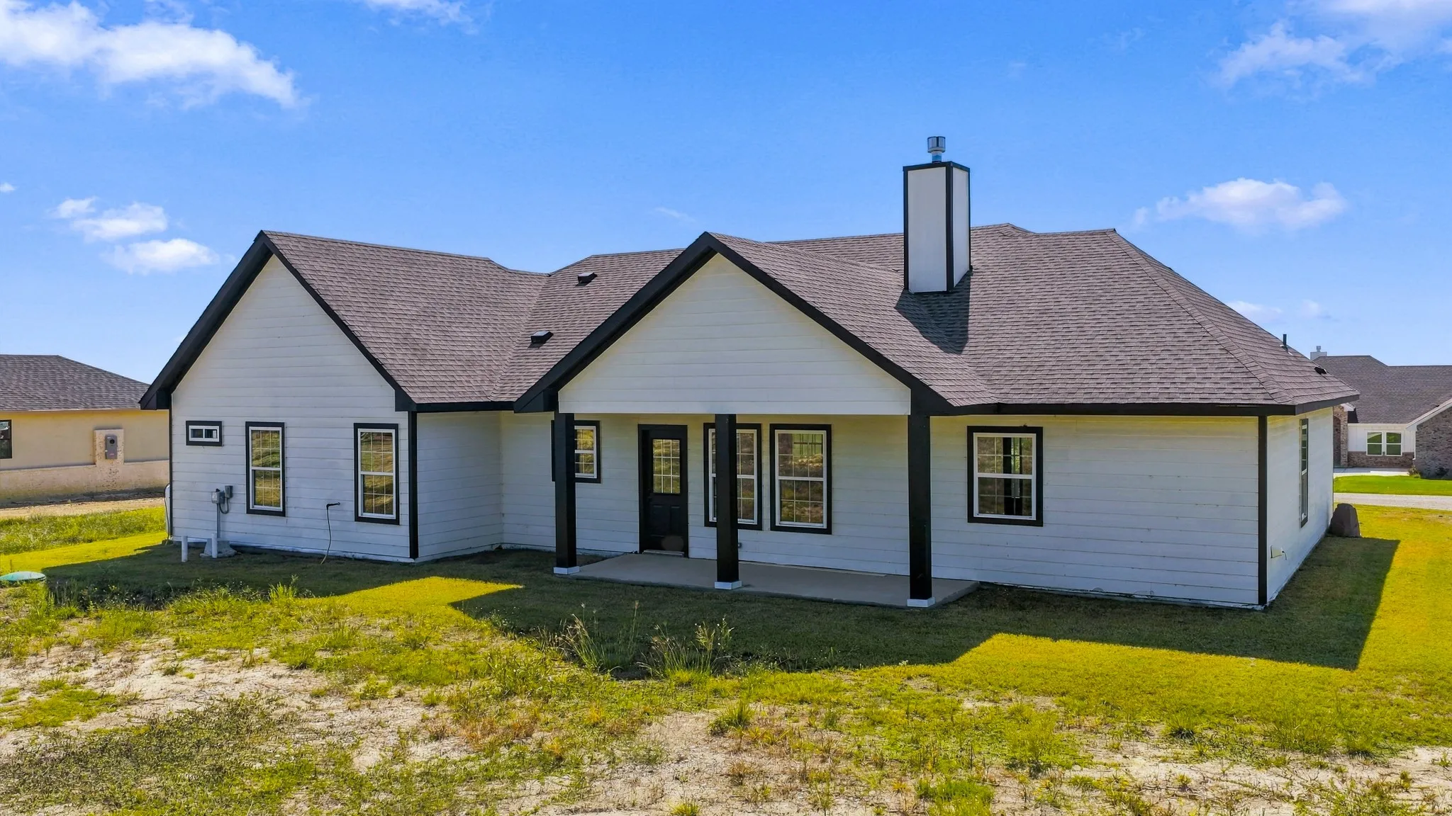 Single-story exterior featuring white horizontal siding, contrasting black trim, an asphalt shingle roof, a covered concrete patio, and a central chimney