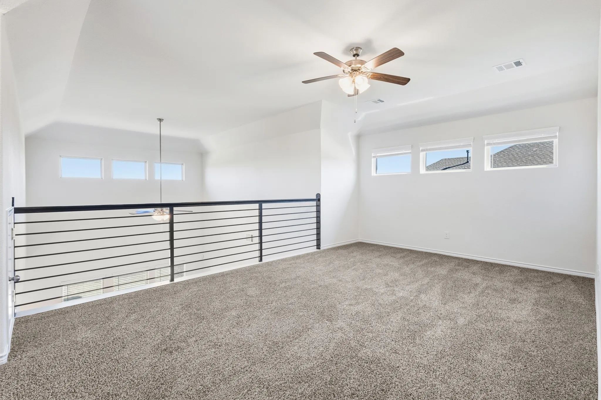 Carpeted loft space featuring multiple windows, a ceiling fan, and a contemporary horizontal bar railing