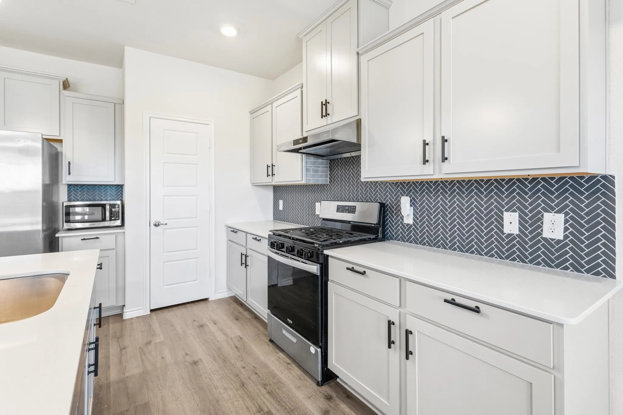 Contemporary kitchen featuring light wood-finish flooring, white cabinetry with black hardware, white countertops, a herringbone tile backsplash, and stainless steel appliances