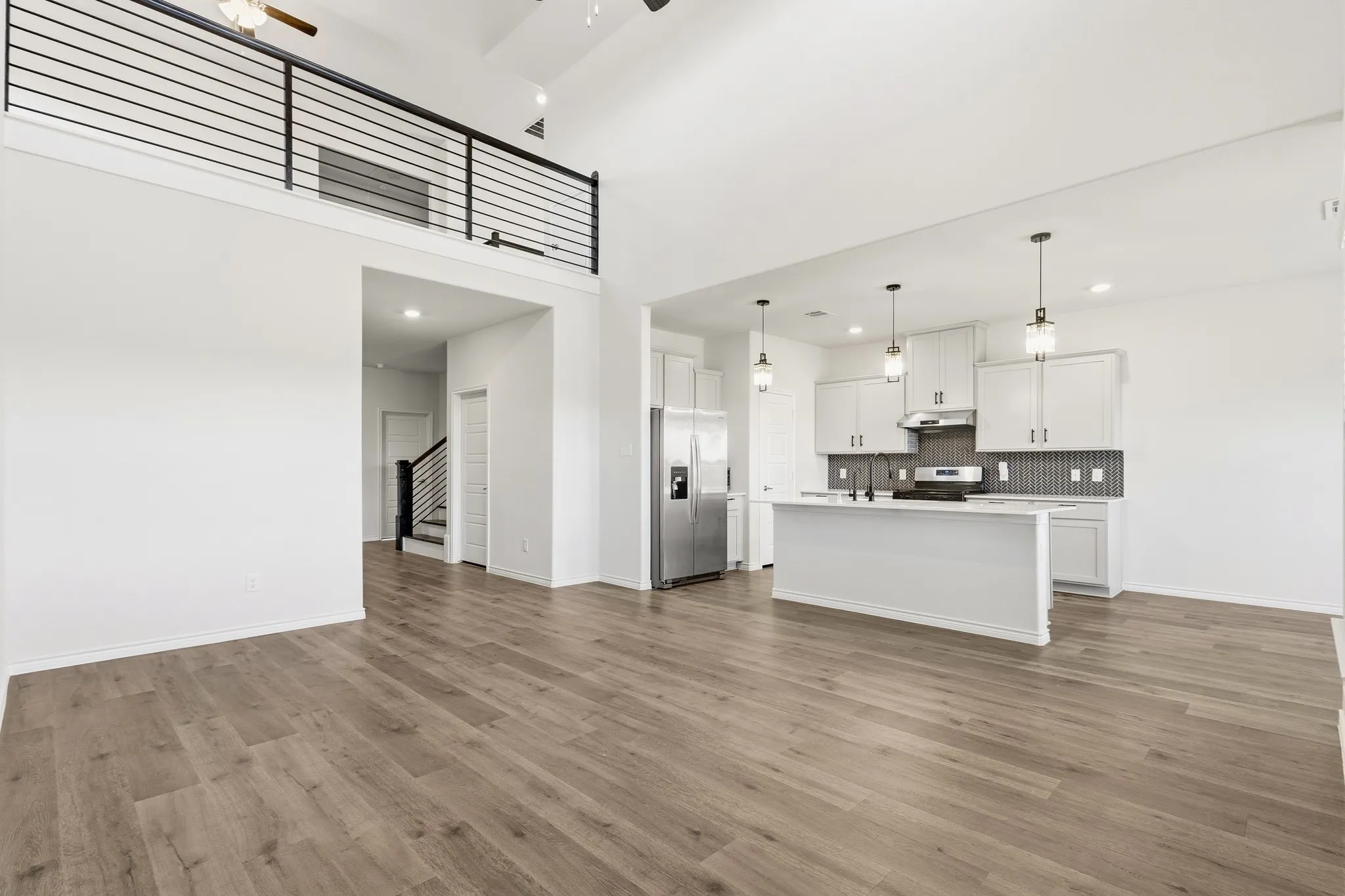 Open-concept living and kitchen area featuring wood-finish flooring throughout, soaring ceilings with pendant lighting, and a modern kitchen with white cabinetry and dark tile backsplash