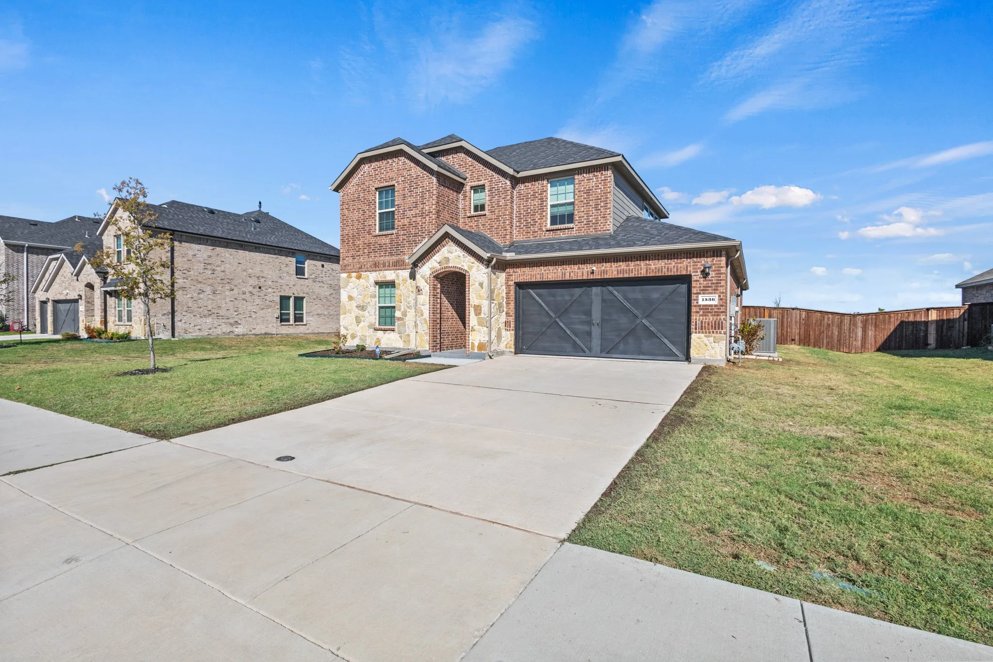 Two-story brick and stone residence featuring a two-car garage with dark paneled doors, established landscaping with mature lawn, and a wide concrete driveway